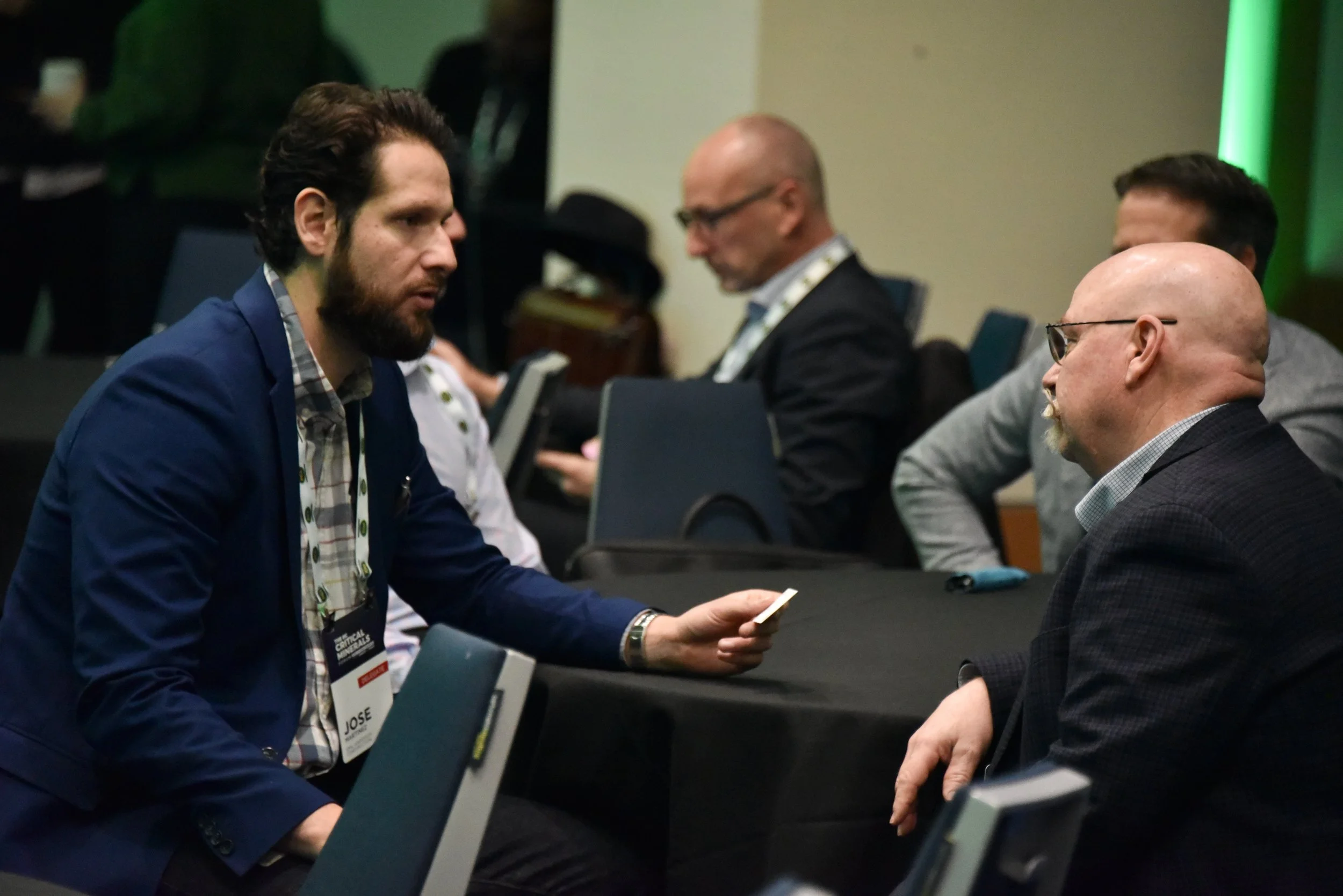 Two men are sitting at a table in a conference room, engaged in a conversation. The man on the left has a beard, dark hair, and is wearing a blue blazer and a checkered shirt, holding a piece of paper. The man on the right is bald, wearing glasses, a