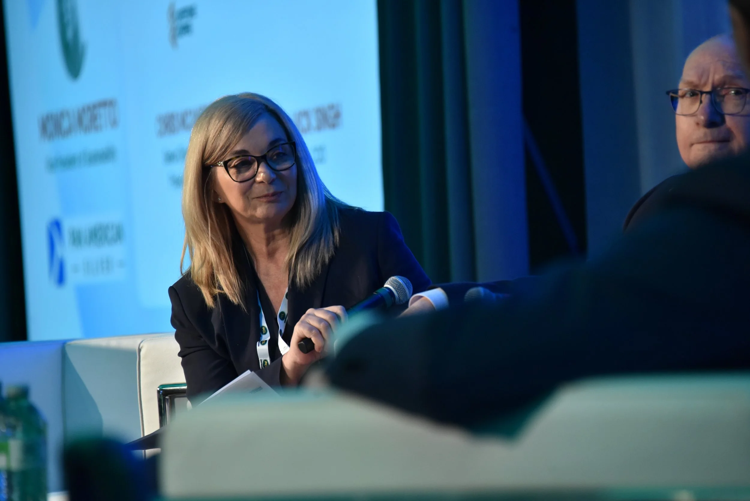 A woman with blonde hair, glasses, and wearing a black blazer, sitting and holding a microphone, during a conference or panel discussion. A man with glasses is partially visible on the right side.