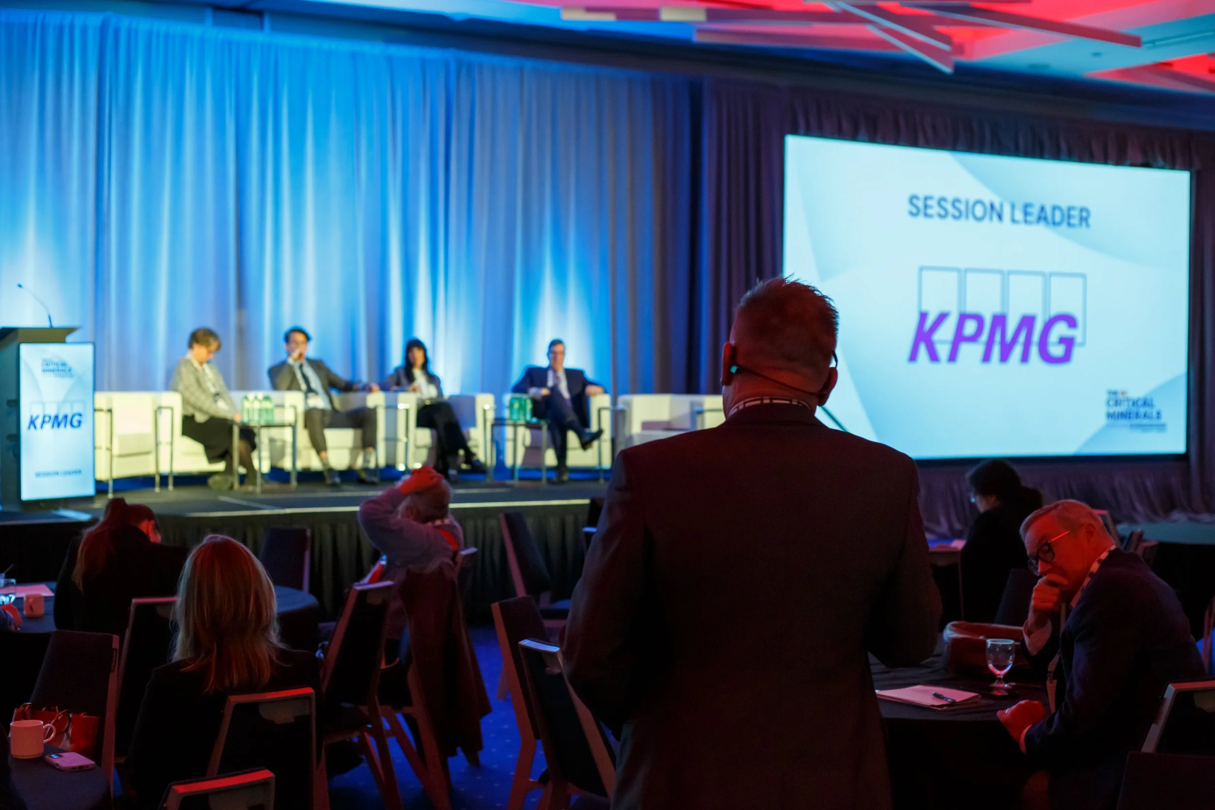 A conference room with a stage where four panelists sit and a large screen that reads 'SESSION LEADER' with the KPMG logo. Audience members are seated and listening, with some taking notes or using their phones.