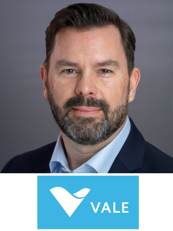 Close-up portrait of a man with short dark hair, beard, wearing a navy blazer and light blue shirt, against a gray background. Below is a blue logo with a stylized check mark and the word 'VALE'.