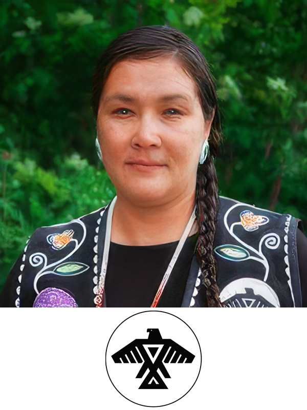 A woman with brown hair in a braid, wearing traditional Indigenous clothing with floral and animal designs, standing outdoors with green foliage in the background, and a black and white emblem of an eagle.»