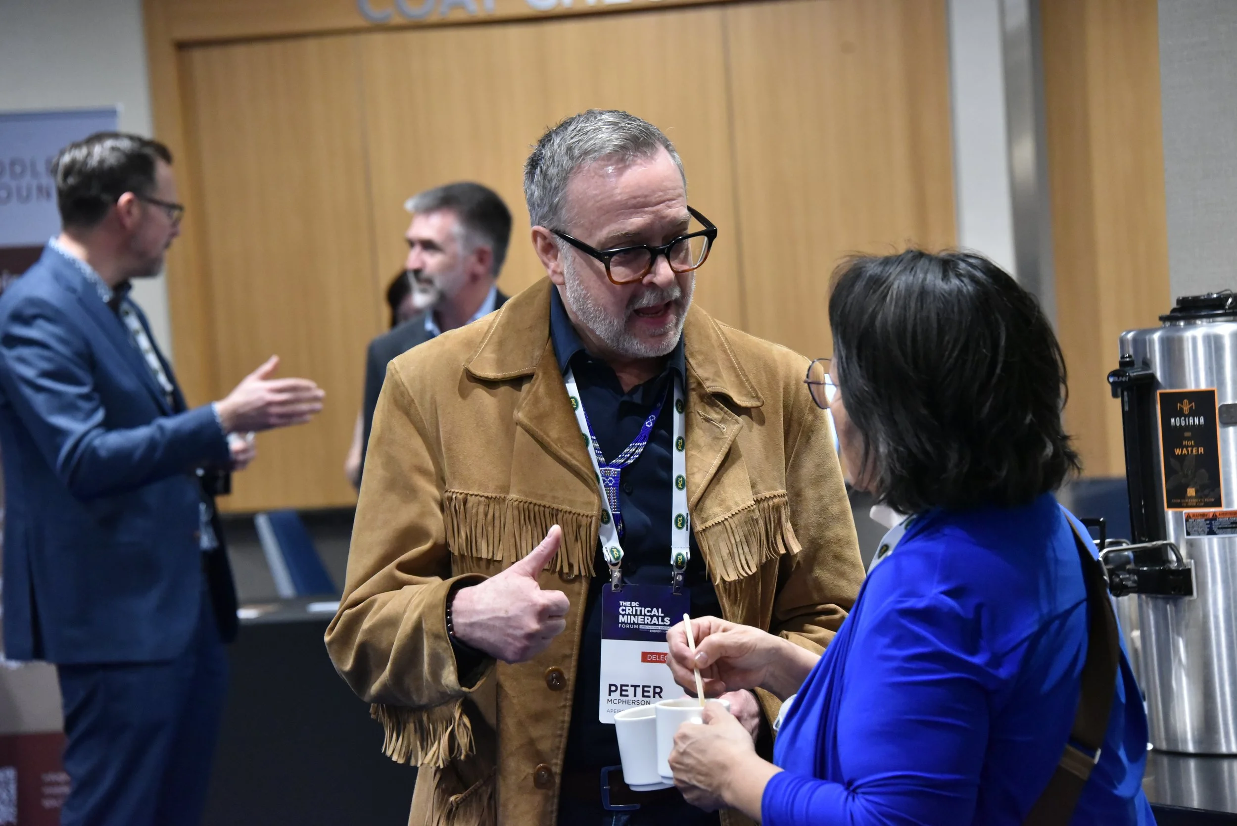 People engaged in conversation at a conference or event, with a focus on a man wearing glasses and a brown fringe jacket talking to a woman in a blue jacket, with others in the background.