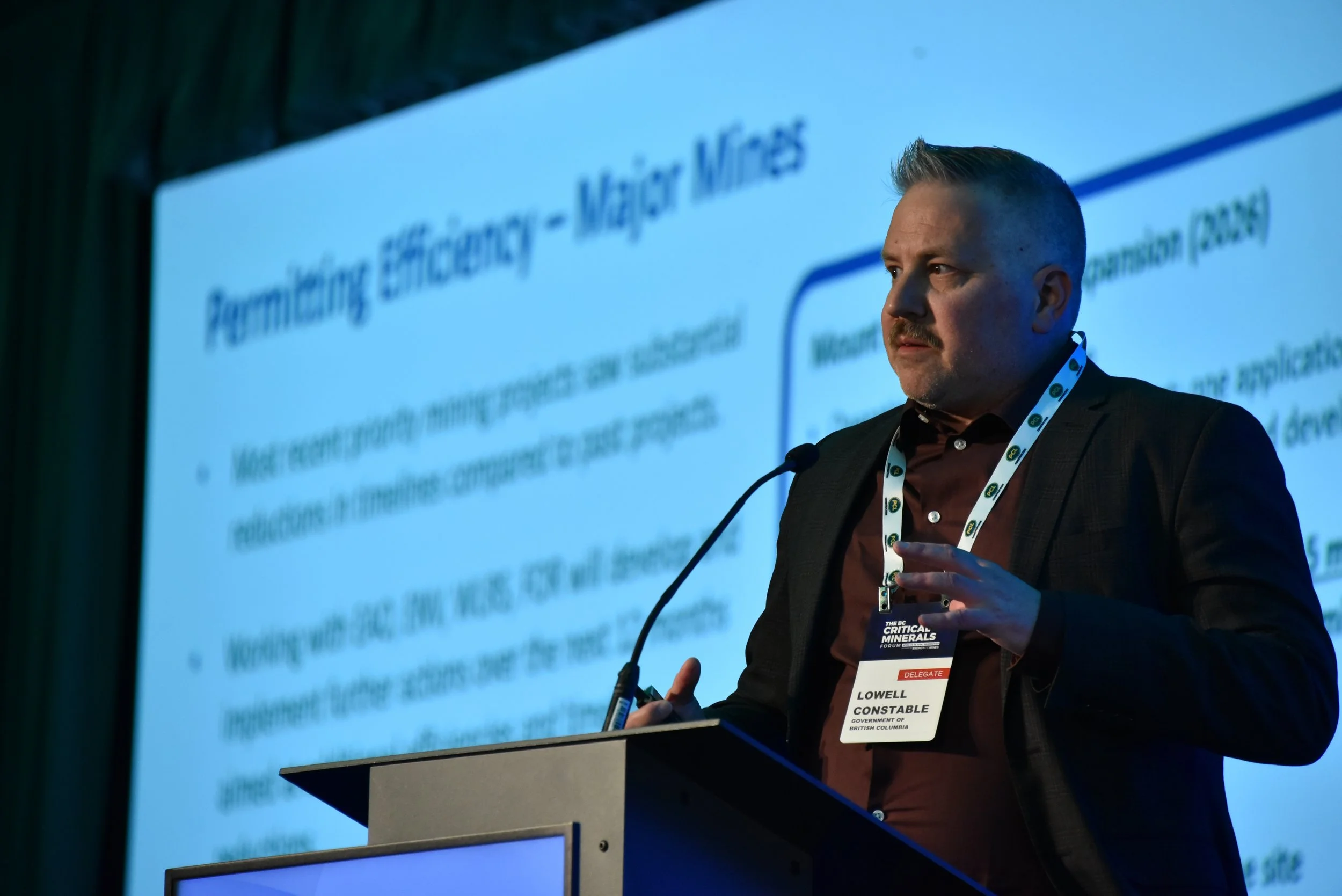 A man in a suit and tie standing at a podium, giving a presentation at a conference. The background features a screen displaying a presentation slide with partially visible text. The man is wearing a conference badge that identifies him as Lowell, a 