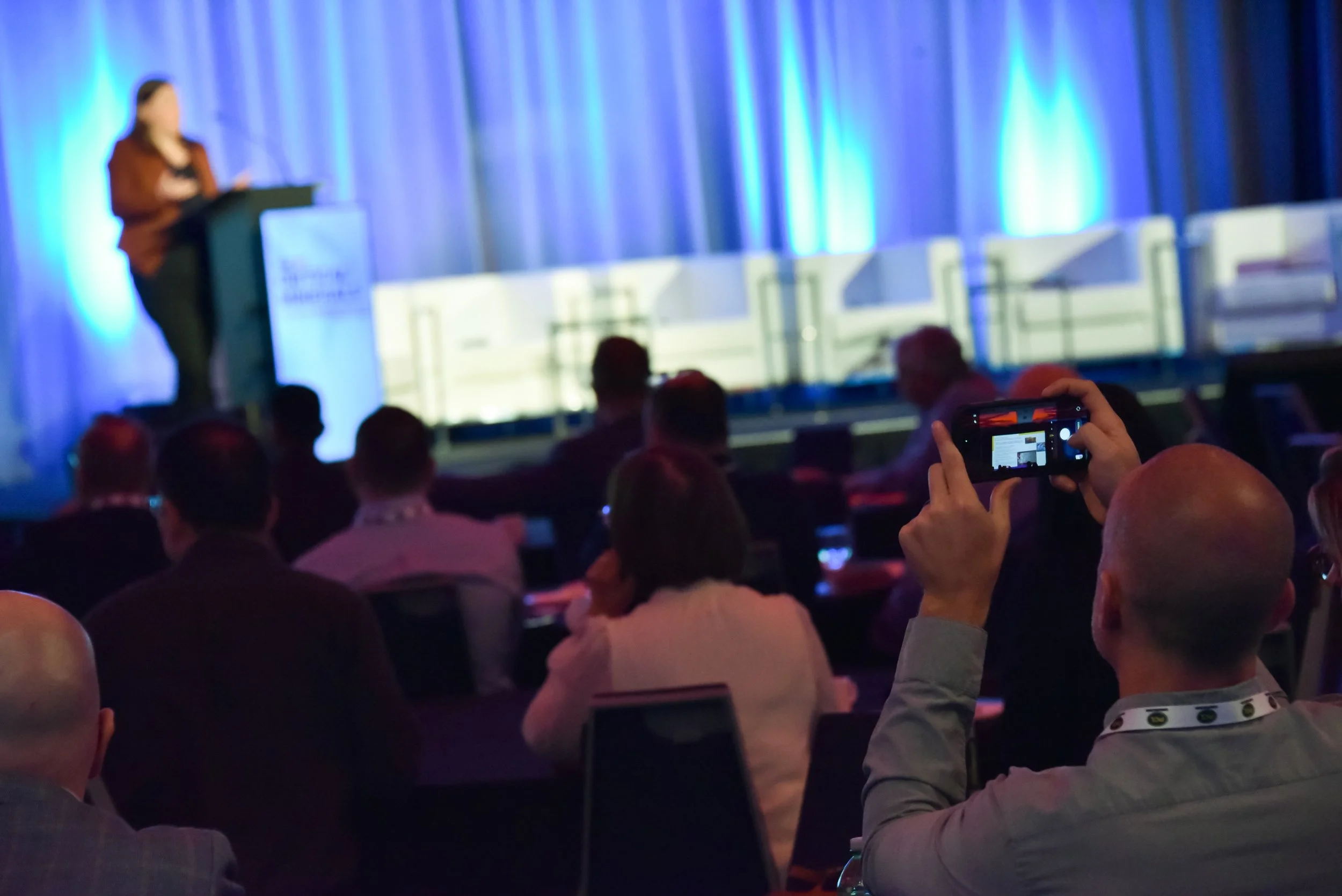 Audience seated at a conference listening to a speaker on stage with a blue-lit curtain backdrop; some audience members are taking photos with their smartphones.