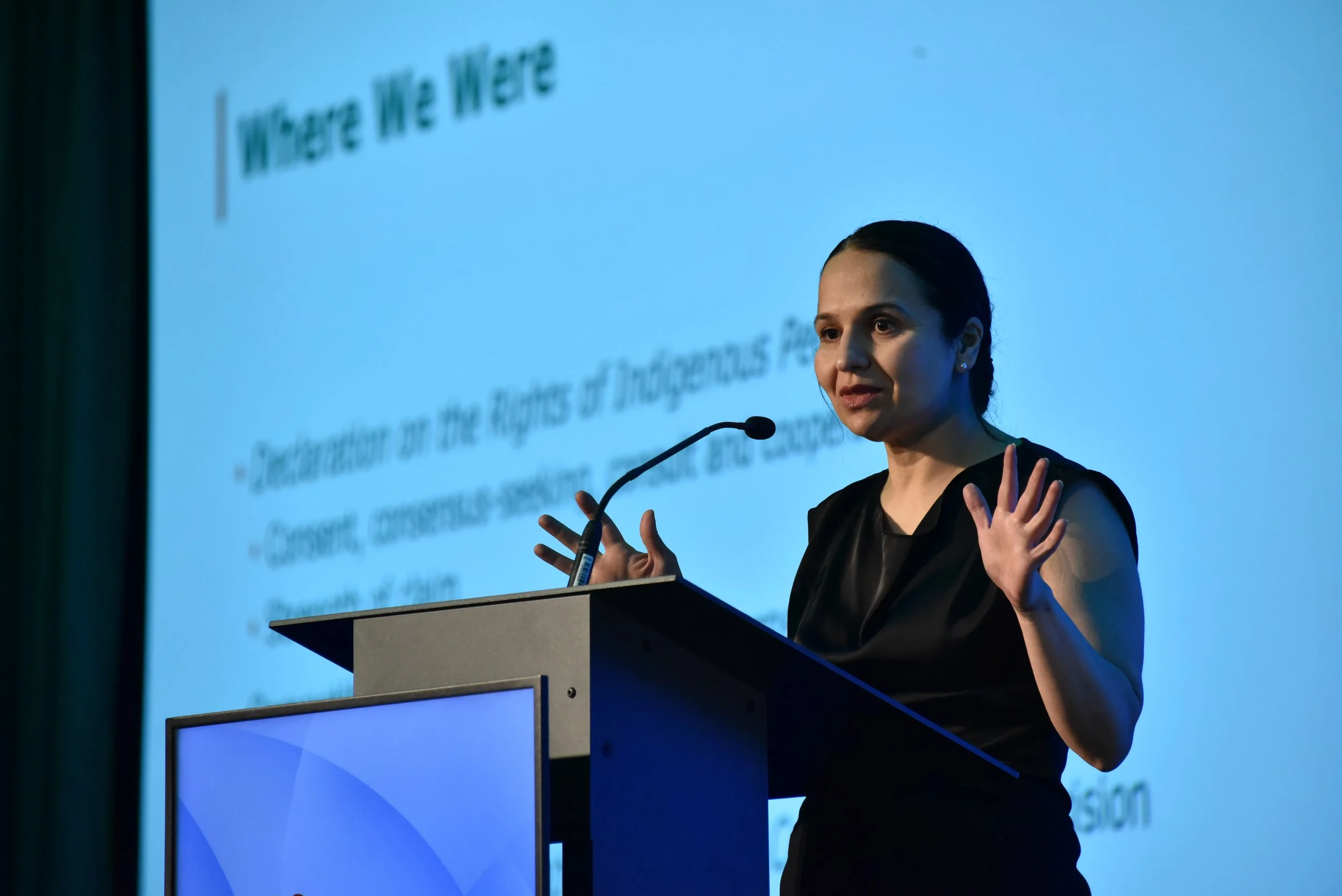 Woman giving a presentation at a podium with a large projection screen behind her displaying the words 'Where We Are'.