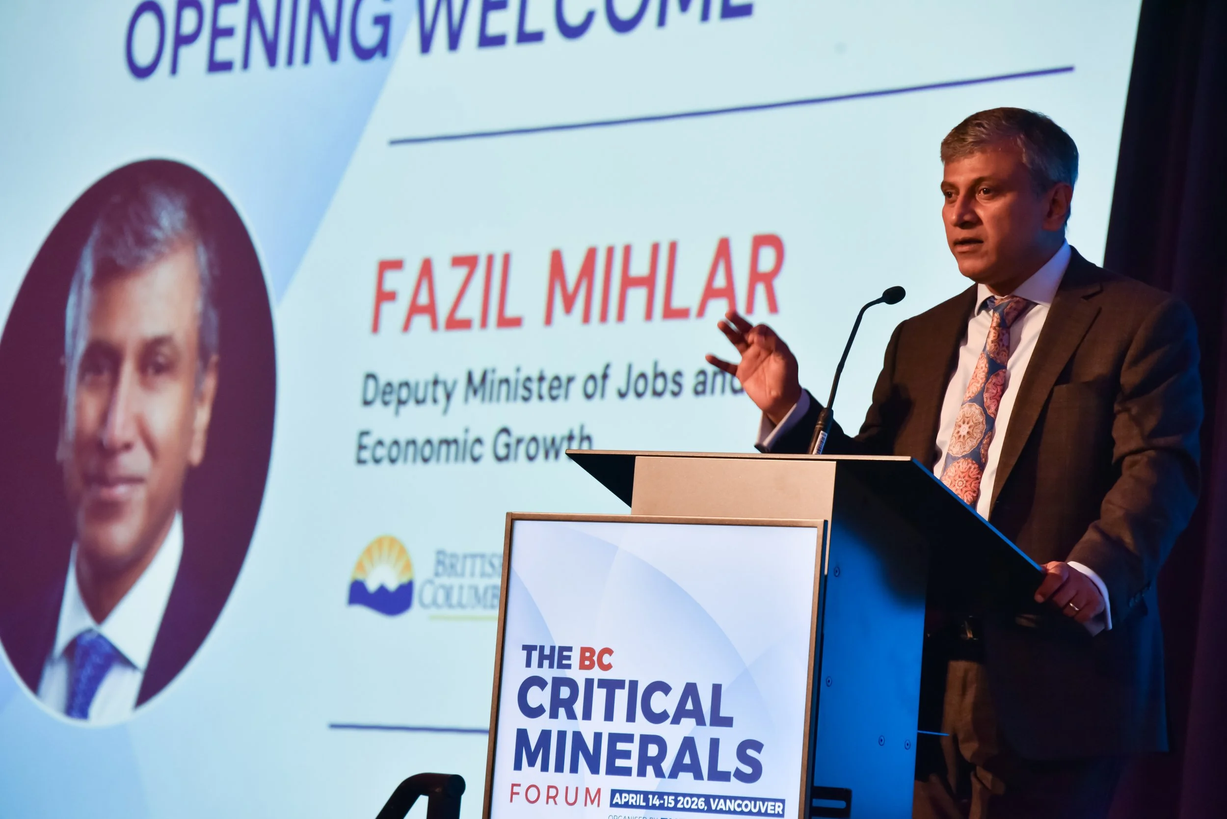 Man in a dark suit and patterned tie giving a presentation at a conference, with a large screen behind him displaying information about a forum on critical minerals scheduled for April 14-15, 2026, in Vancouver. Part of a circular photo of a woman is