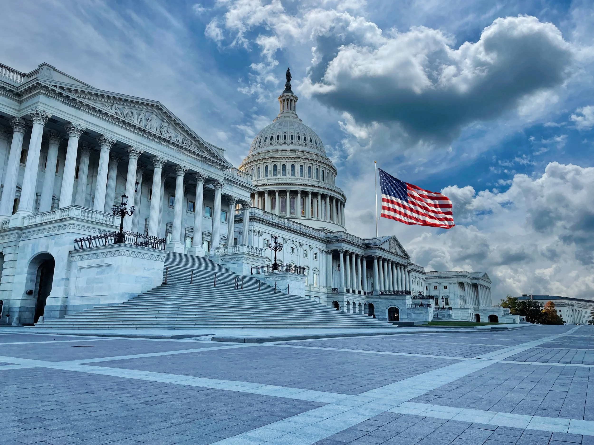 The image shows the United States Capitol building with its iconic white dome and tall columns. There is an American flag flying in front of the building, and the sky above is partly cloudy with a mix of blue sky and white clouds. The area in front of the building has wide steps and a paved plaza.