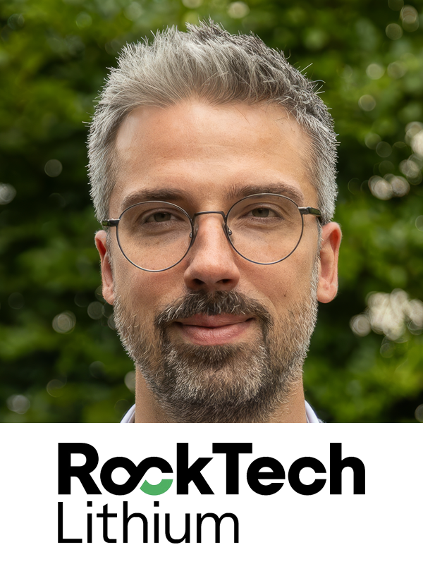 A man with gray hair and a beard wearing round glasses, standing outdoors with green foliage in the background. The bottom of the image features the logo 'RockTech Lithium'.