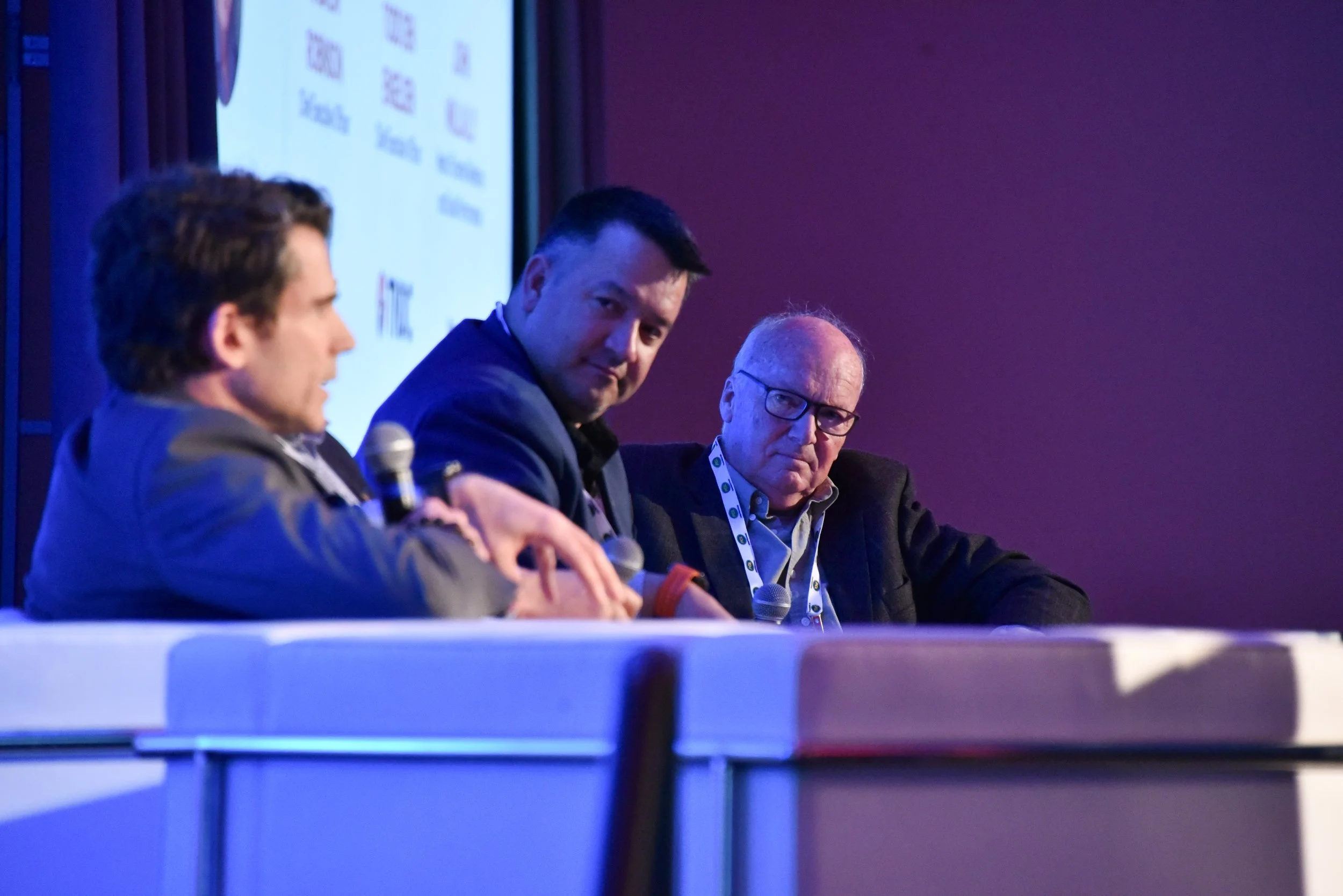Four men seated at a panel discussion or conference, with microphones in hand. The background includes a presentation screen with visible text and logos, and purple lighting.