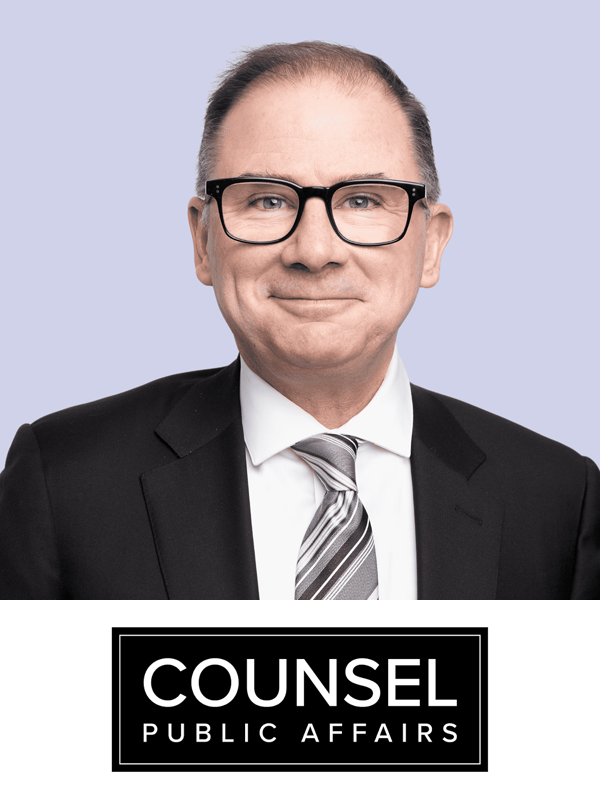 A professional man wearing glasses, a black suit, white shirt, and striped tie, posing for a portrait against a light purple background, with a "Counsel Public Affairs" logo at the bottom of the image.