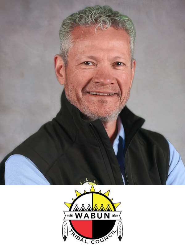 A middle-aged man with gray hair and a beard, wearing a light blue shirt and a black vest with a logo, smiling for a portrait against a gray background, with the Wabun Tribal Council logo at the bottom.