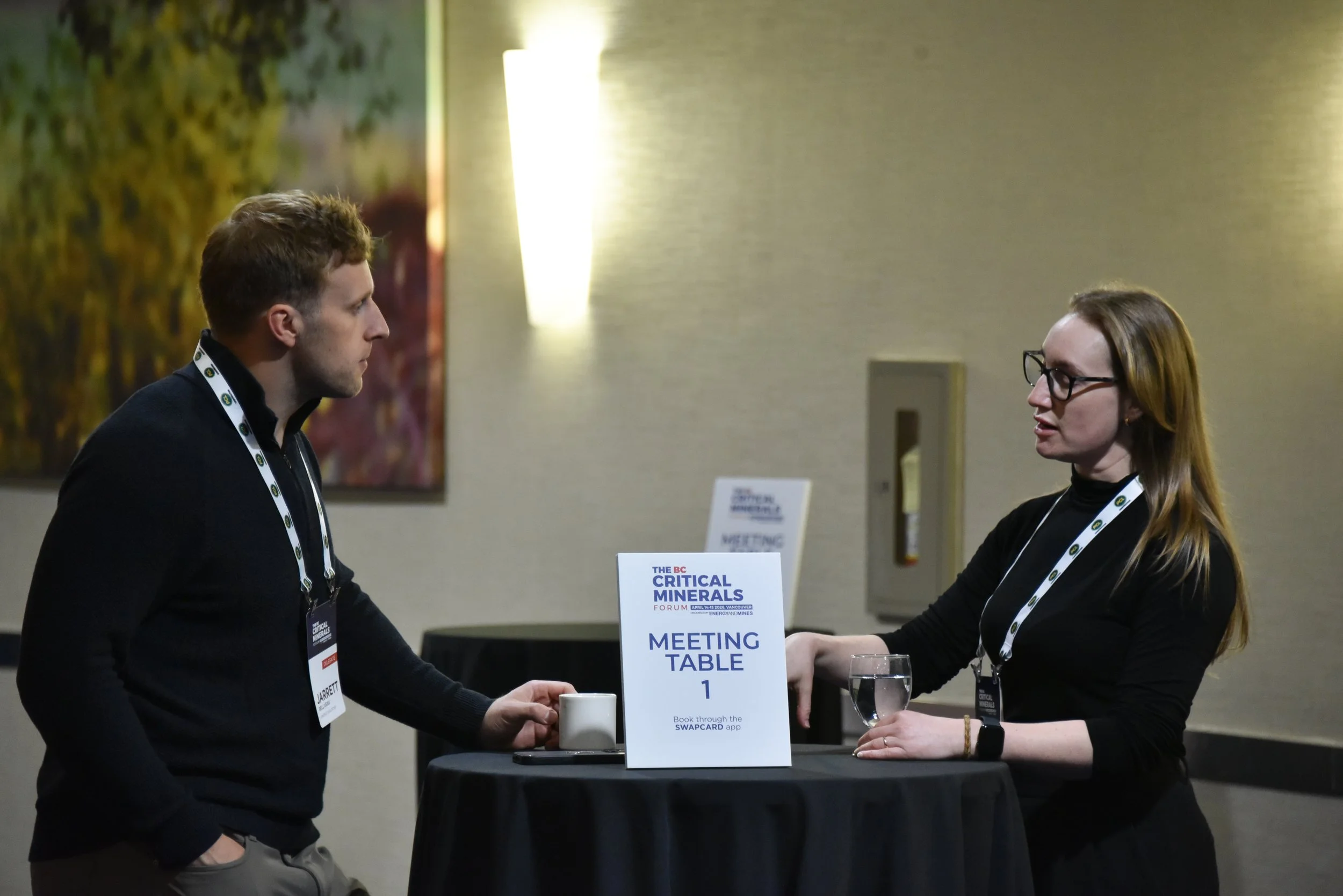 Two individuals engaging in conversation at a meeting table during a conference, with signs indicating it is the BC Critical Minerals Forum.