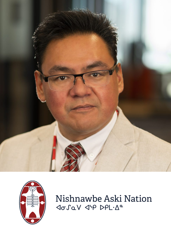 Portrait of a man with glasses wearing a beige suit, white shirt, and red tie, in an indoor setting with blurred background. Includes a logo and text reading 'Nishnawbe Aski Nation' and its native language translation.