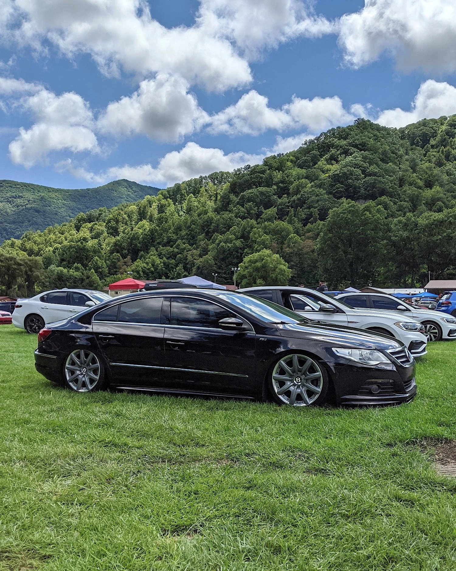 Black Volkswagen CC lowered on air suspension with Bentley wheels parked on grass at a mountain car show