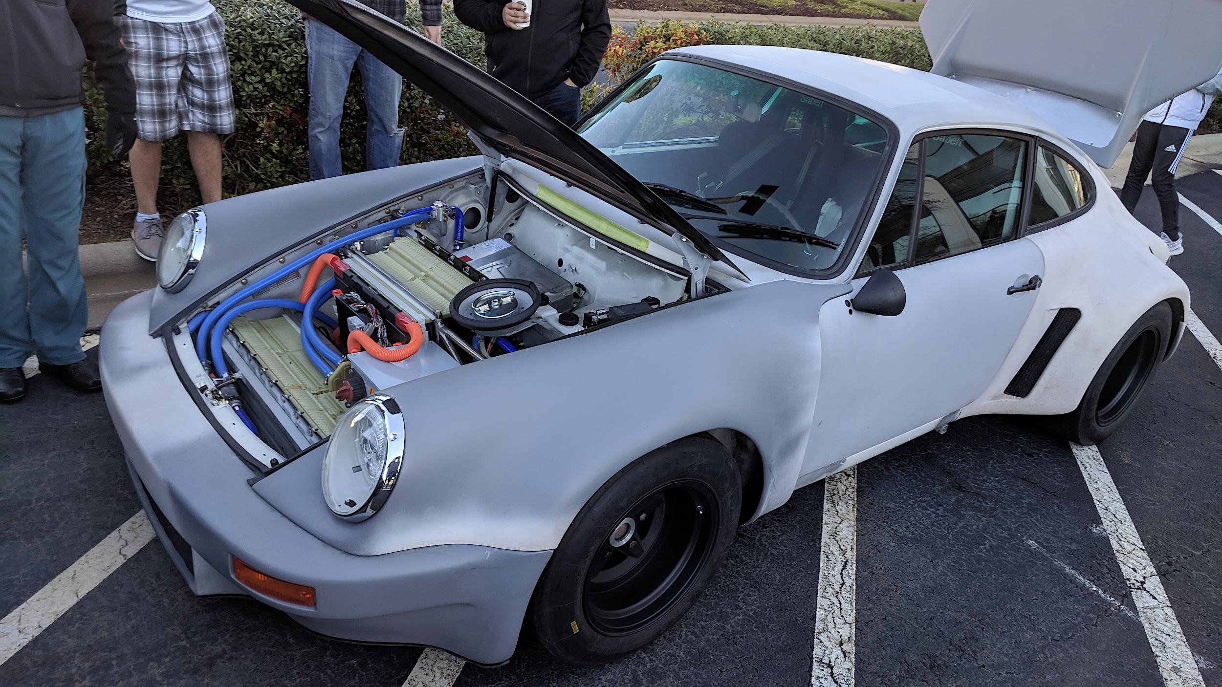 Gray primered widebody Porsche 911 with a Tesla electric powertrain, both lids open at a morning cars and coffee