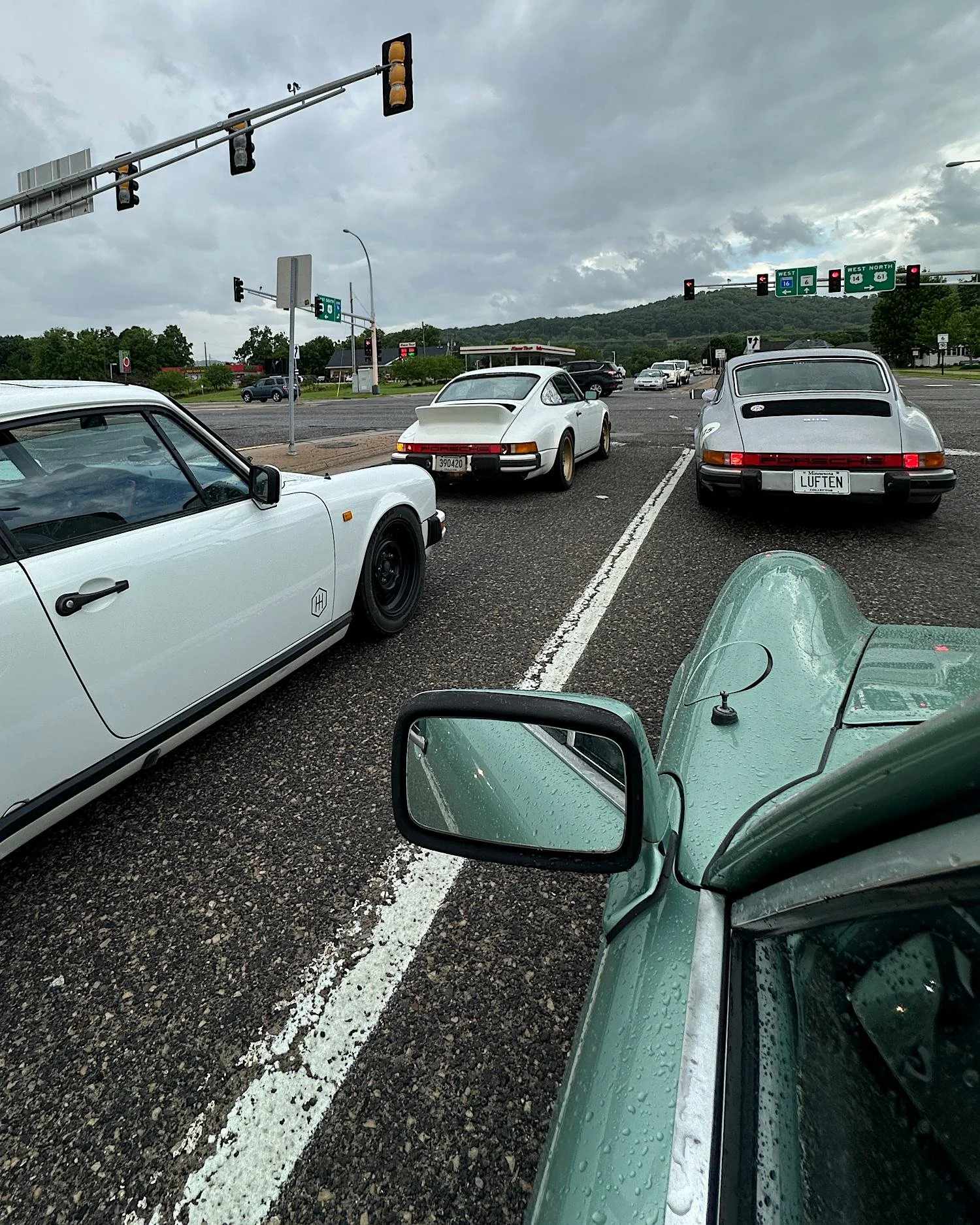 Driver's view over the wet fender of an ice green 911 stopped at a traffic light, with three white and silver 911s waiting in the lanes ahead