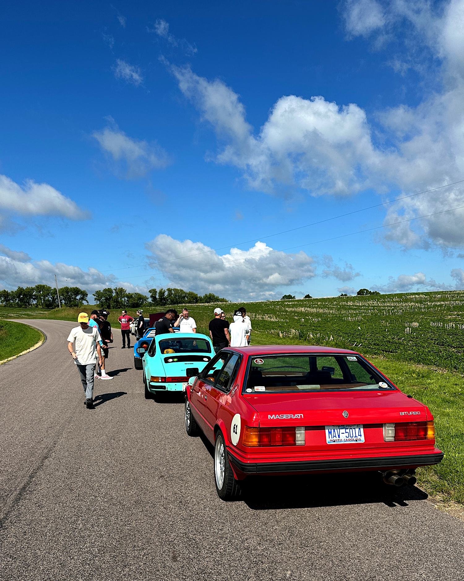 A Maserati Biturbo, turquoise Porsche 911, and Lancia pulled onto the shoulder of a country road beside a farm field, with drivers and passengers standing around talking.