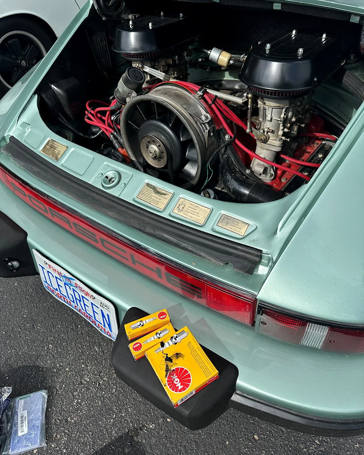 Open engine lid of an ice green 1977 Porsche 911S with twin carburetors and red plug wires, new spark plugs in open boxes on the bumper