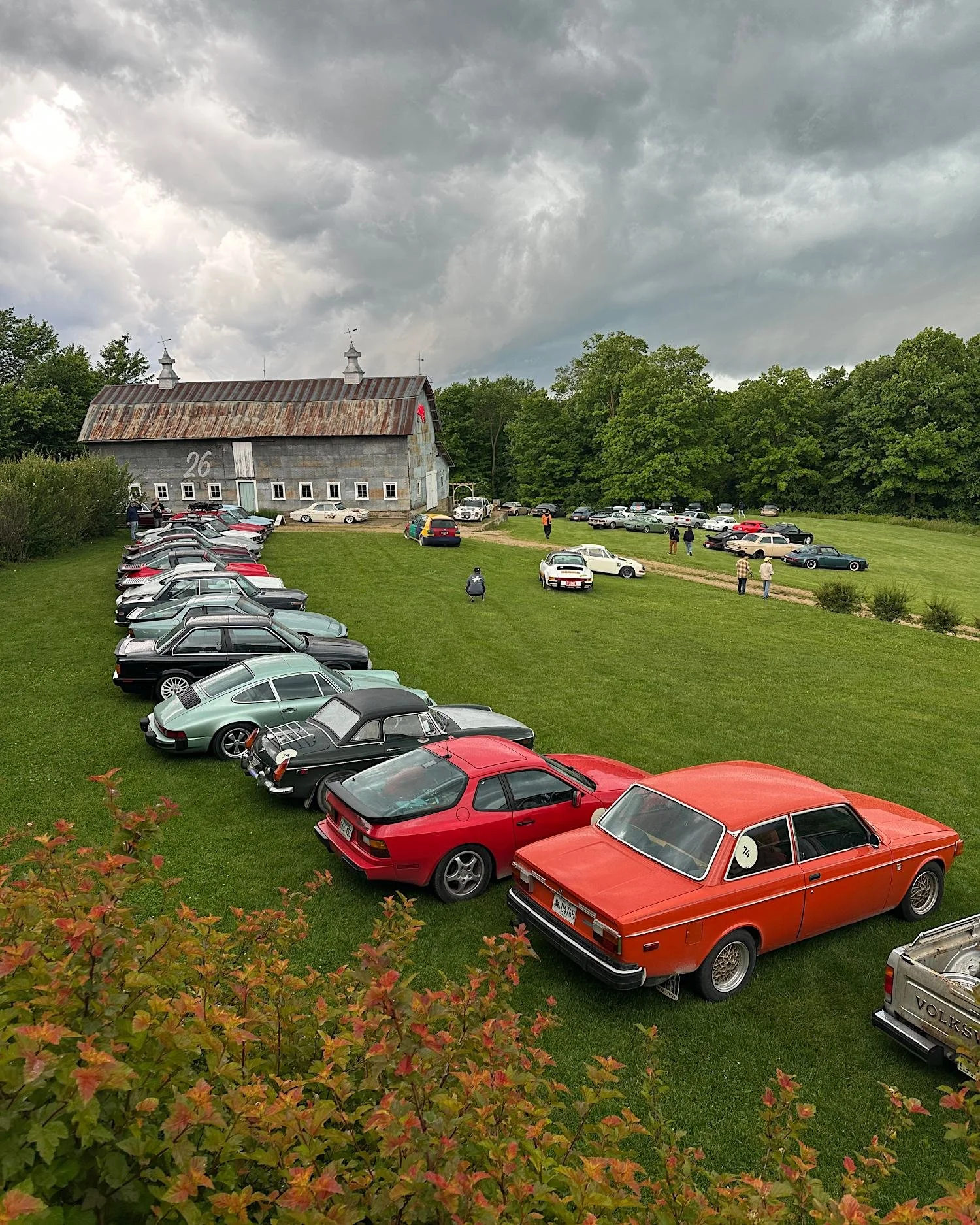 Rows of vintage sports cars parked in a grass field beside a weathered tin-roofed barn under a dark storm sky.