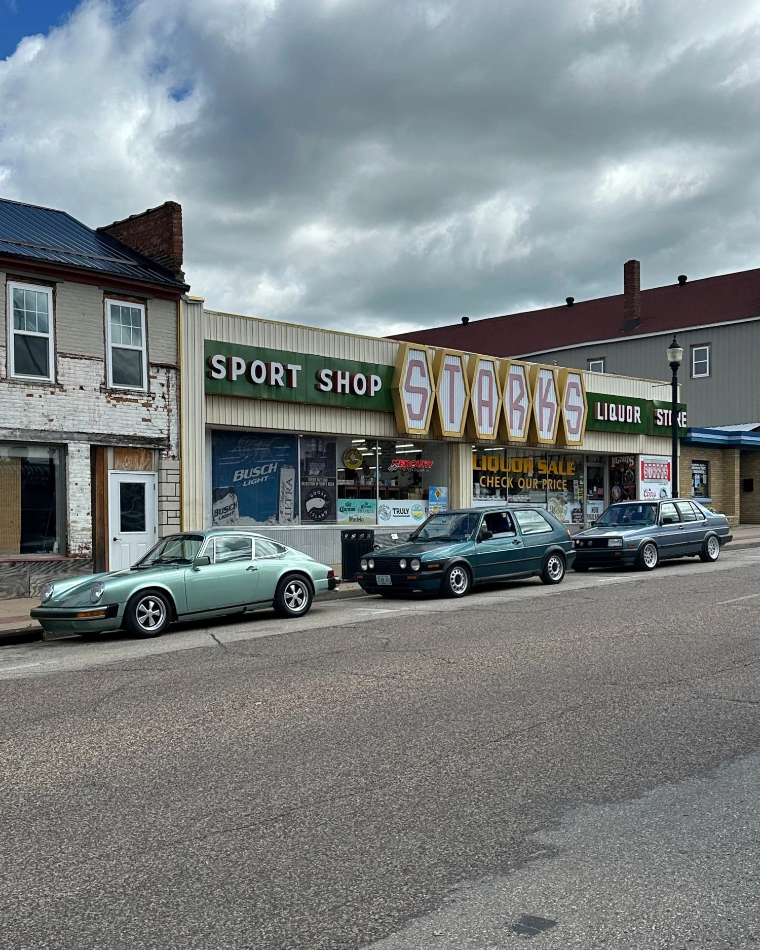 An ice green 911 parked at the curb on a small-town main street with two Mk2 Volkswagens, storefronts and a cloudy sky behind