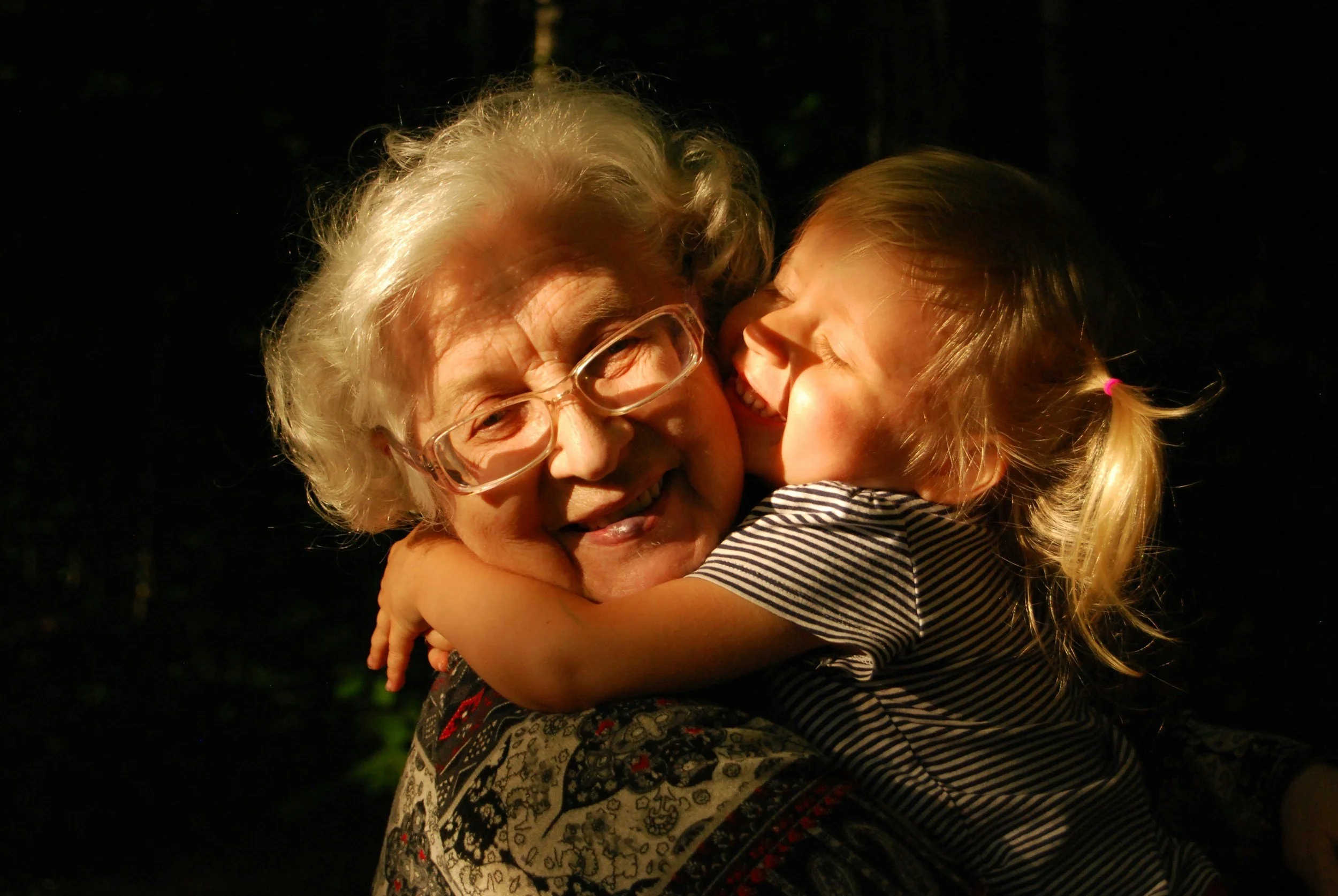 An elderly woman with glasses and white hair embracing a young girl with blonde hair in a ponytail, both smiling and happy in a dark outdoor setting.