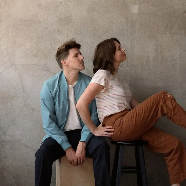 A man and woman sitting against a textured wall, with the woman sitting on a stool. The man is sitting on a box, and the woman in front of him has her back against his chest, while he is leaning slightly backward. She is looking up and to the right, 