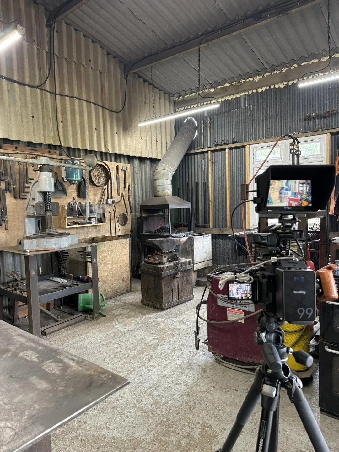 A workshop with metal tools hanging on a wooden board, a forge with a chimney, a workbench, and a camera on a tripod filming the scene.