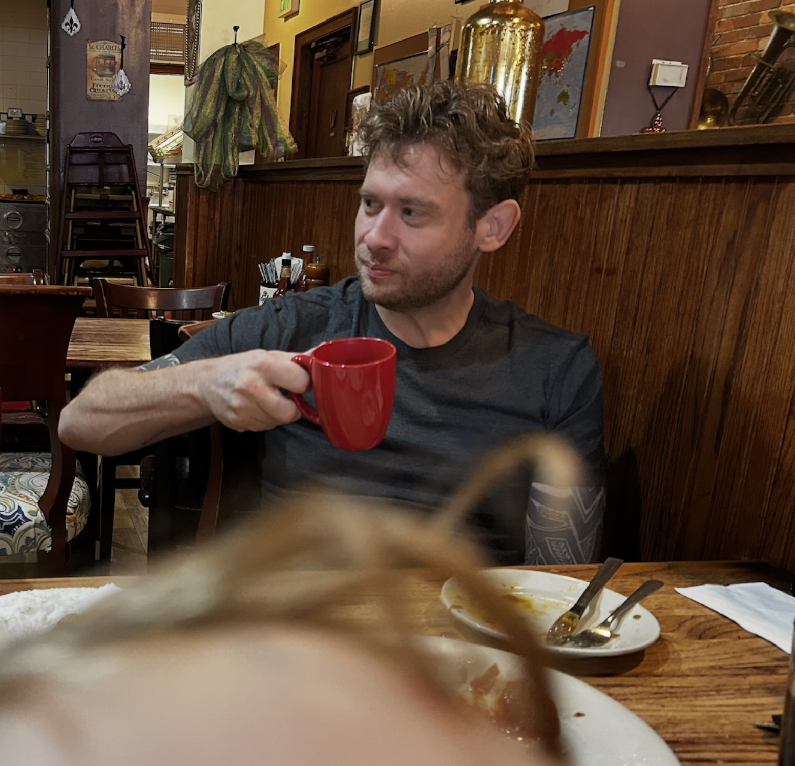 Tom Chalk, creative director of Chalk Outlines Film, sitting at a wooden table in a cozy restaurant holding a red mug. The table has a plate with some remaining food and utensils. The background features wall art, a map, and warm decor.