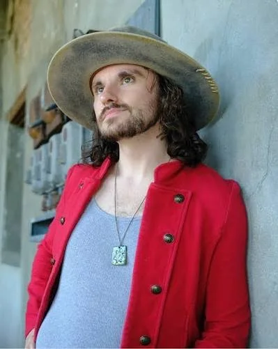 A man with long dark hair and a beard wearing a wide-brimmed hat, a red jacket, and a gray shirt, leaning against a concrete wall.