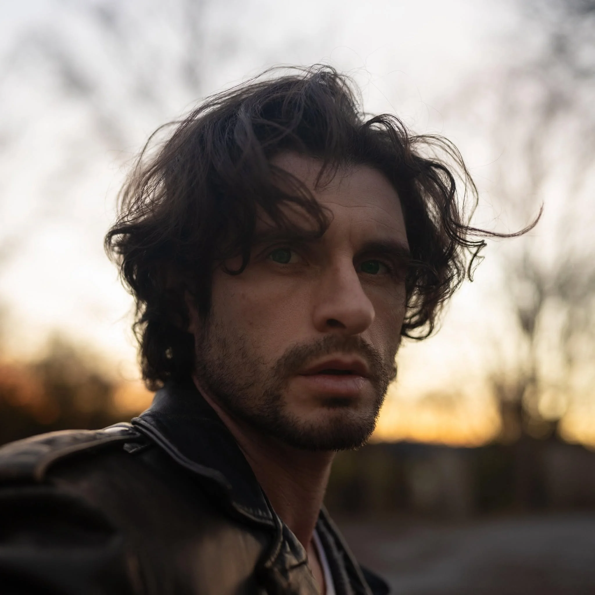Close-up of a man's face with dark curly hair and light skin, outdoors during daytime with blurred trees in the background.