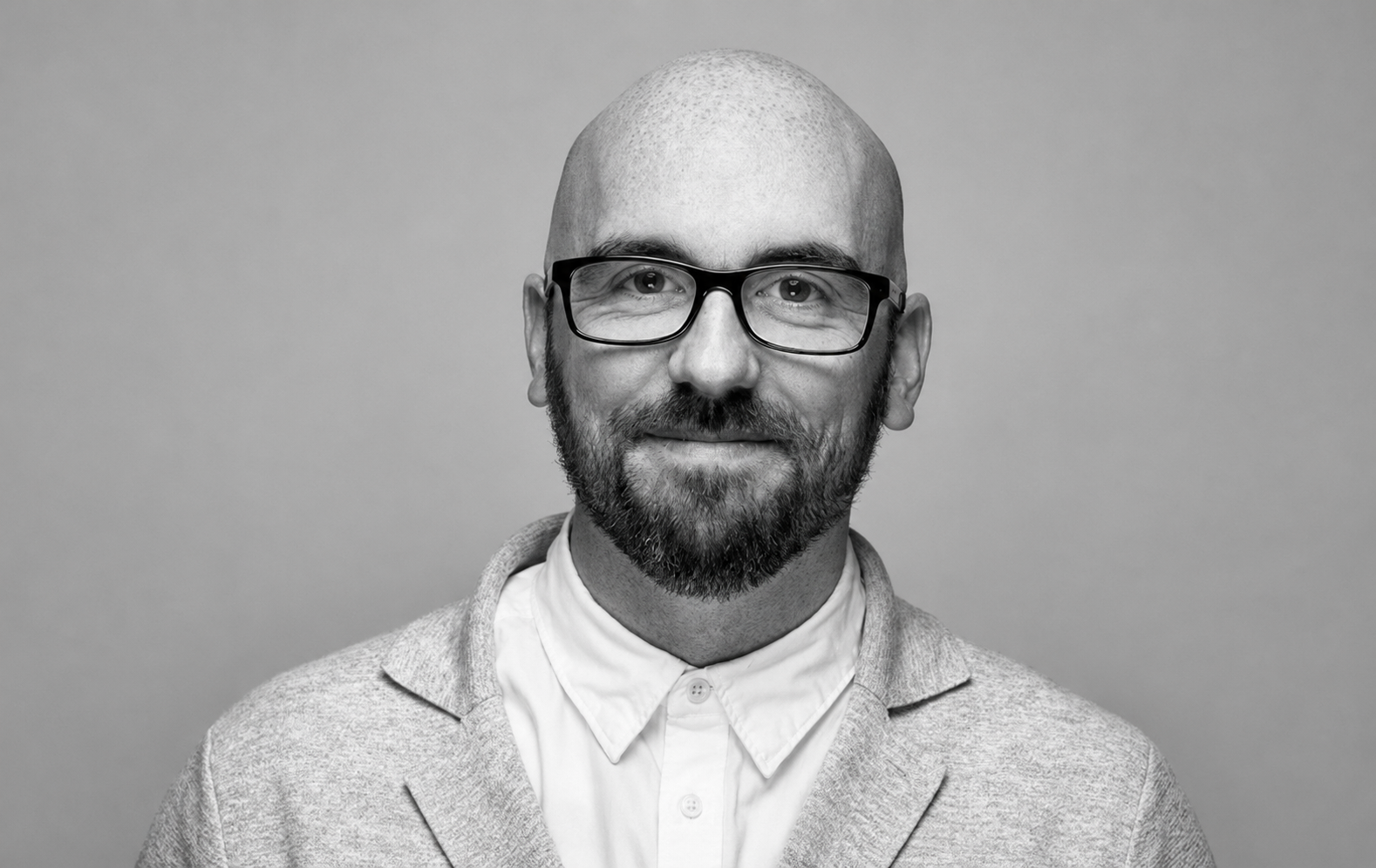 Black and white portrait of a man with glasses, a beard, and a bald head, smiling at the camera, wearing a collared shirt and a sweater, against a plain background.