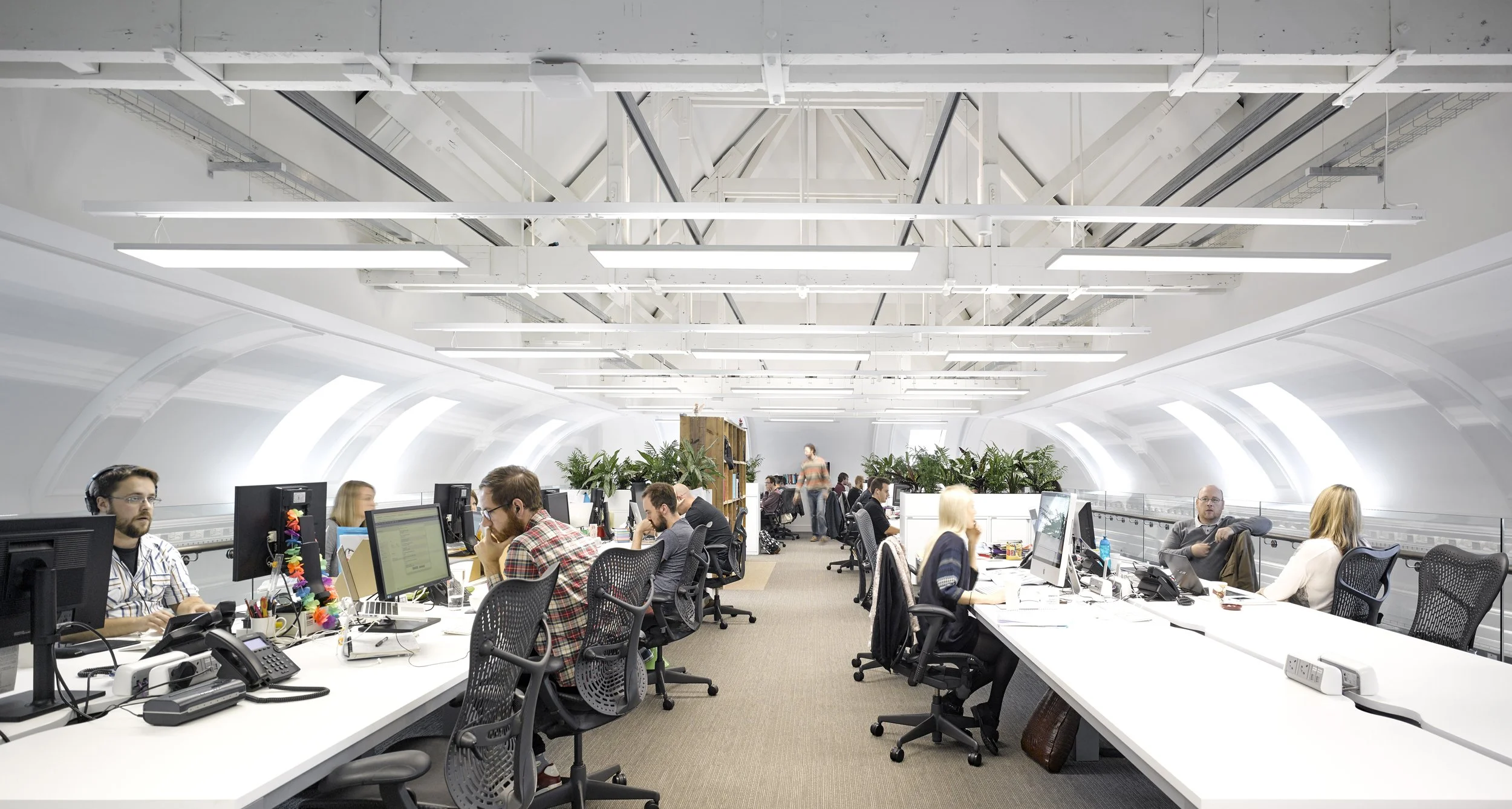 An open office space with many people working at desks with computers, in a modern room with a curved ceiling and bright lighting.