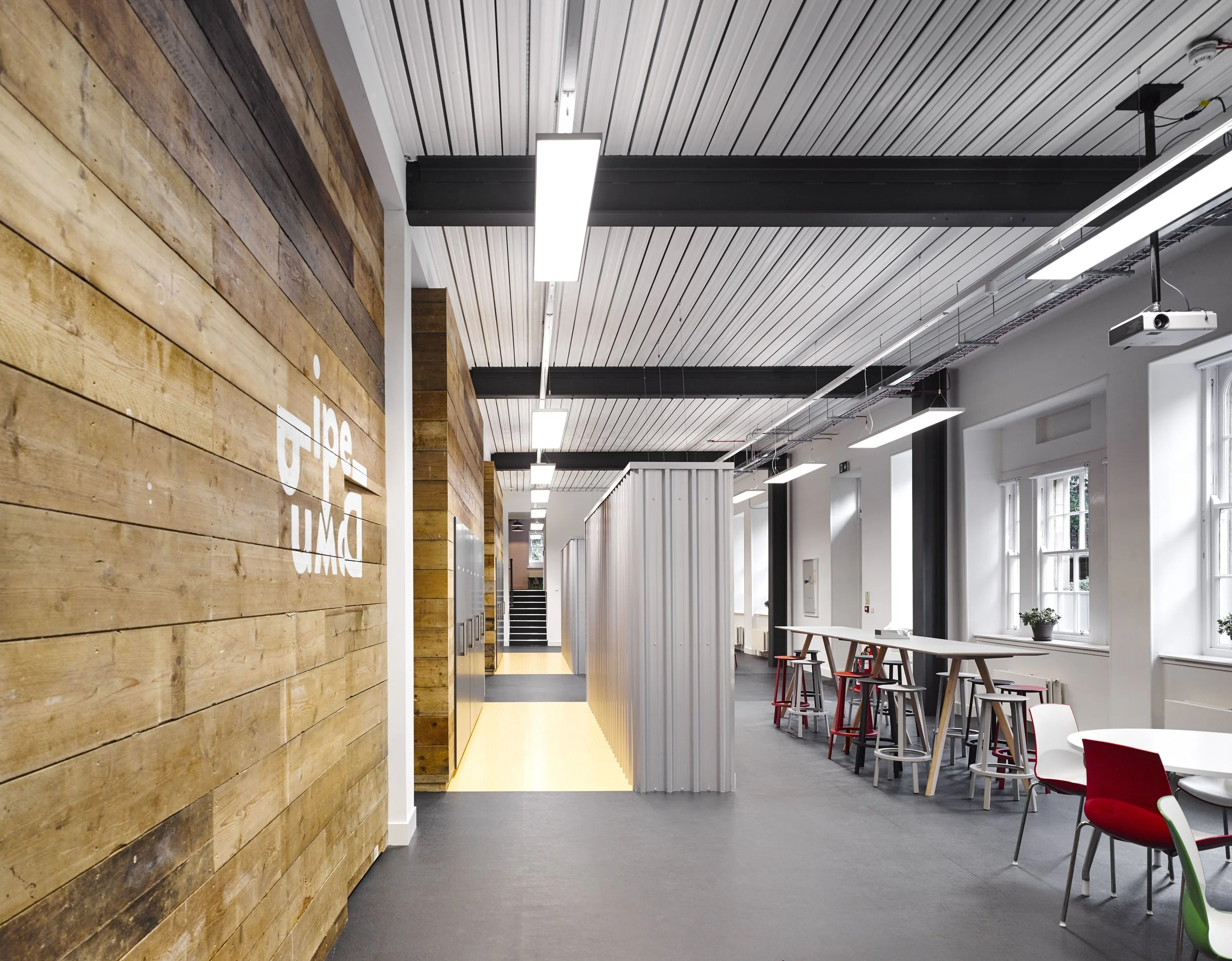 Modern office interior with wooden wall, white ceiling with linear lights, and seating area with tables and chairs.