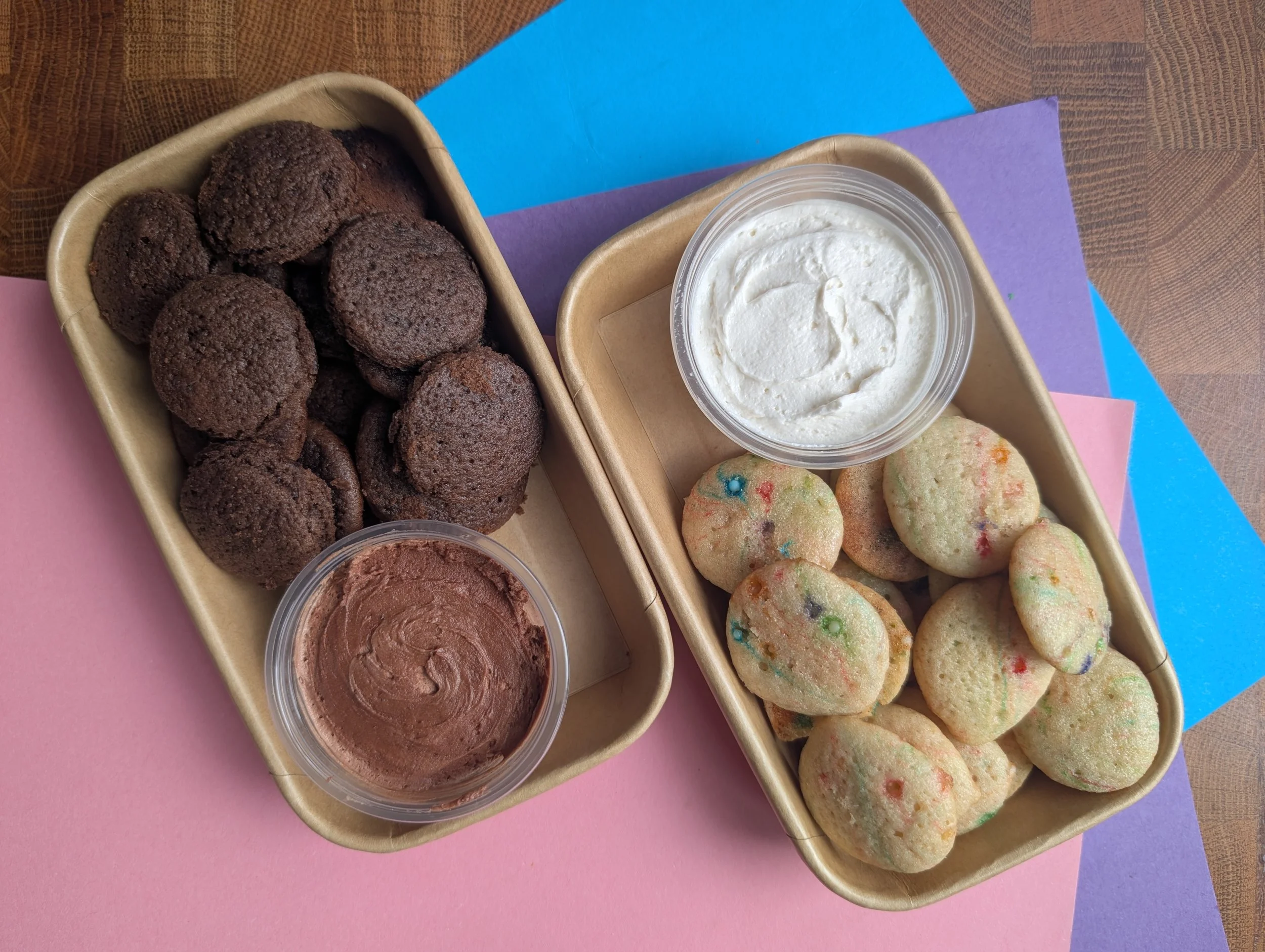 Trays filled with cookies and frosting. One tray has chocolate cookies, the other tray has sugar cookies with colorful sprinkles. There are small containers of frosting, one chocolate and one vanilla, on the trays. Homemade dunkaroos. cookies and dip