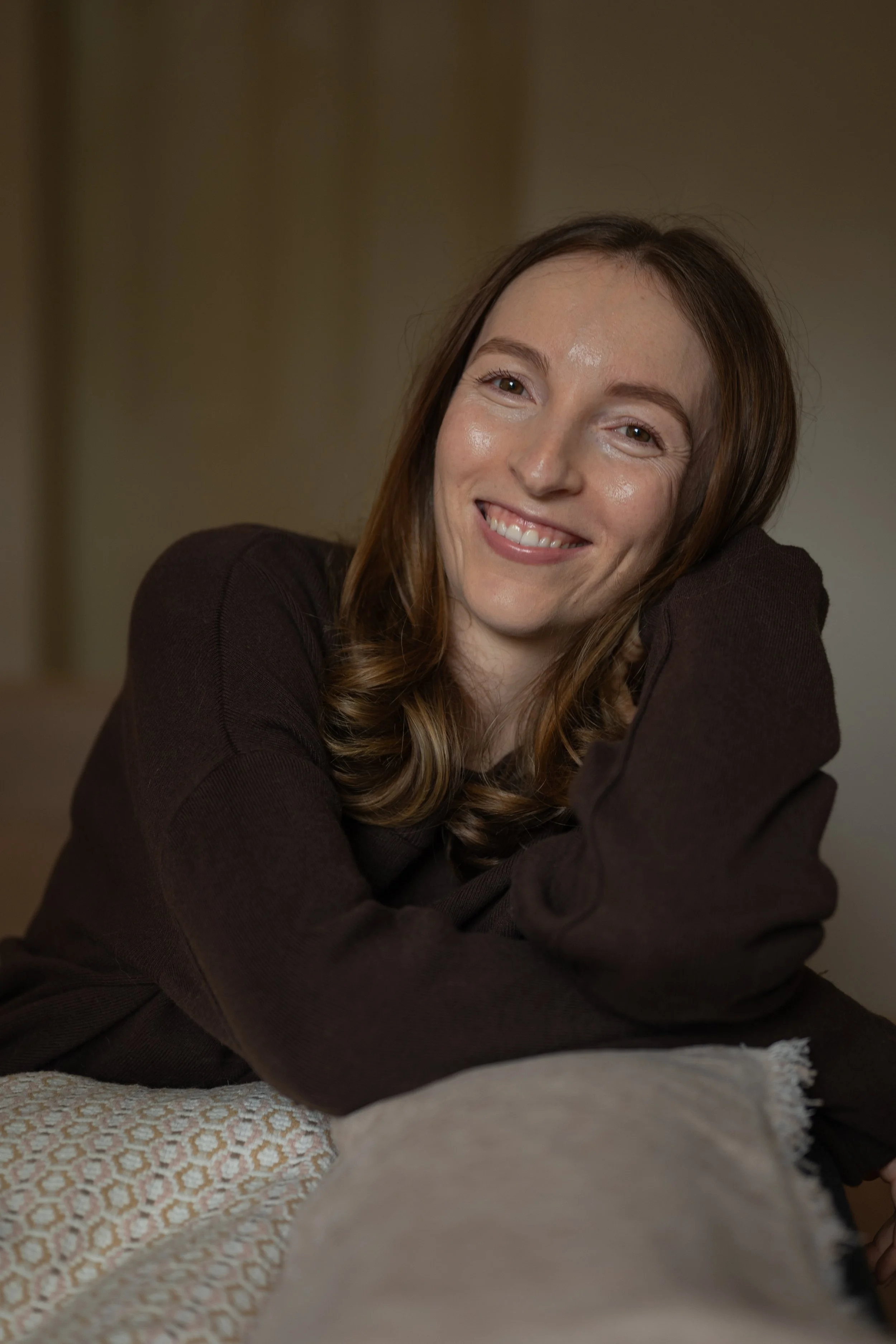 A woman with long brown hair, smiling and resting her face on her hand, wearing a dark brown sweater, sitting at a table with a textured tablecloth.