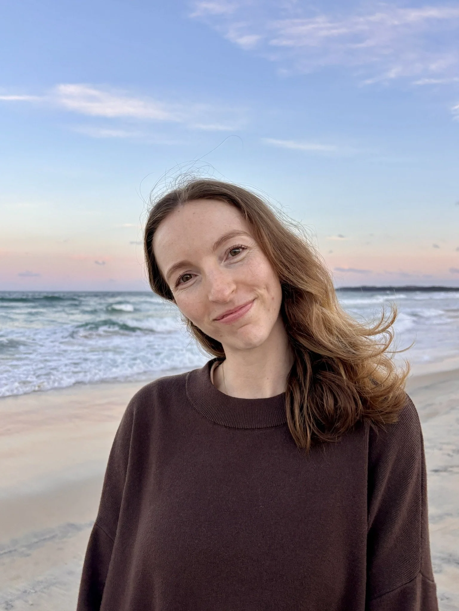 A woman with long, wavy red hair smiling at the camera at the beach during sunset, with the ocean and sky in the background.