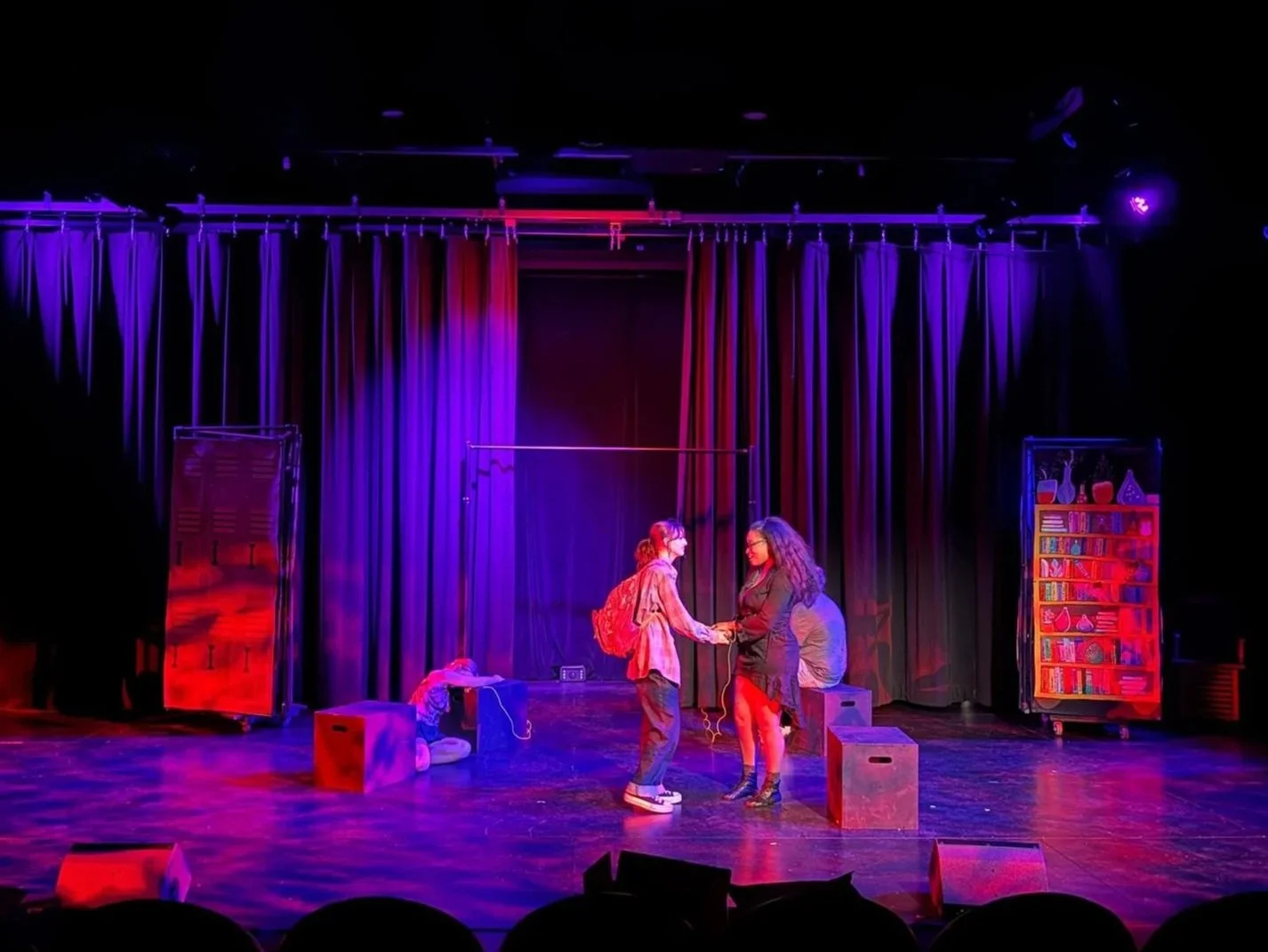 Two women shake hands on a stage with purple curtains, bookshelves, and stage props in the background, illuminated by colored stage lights.
