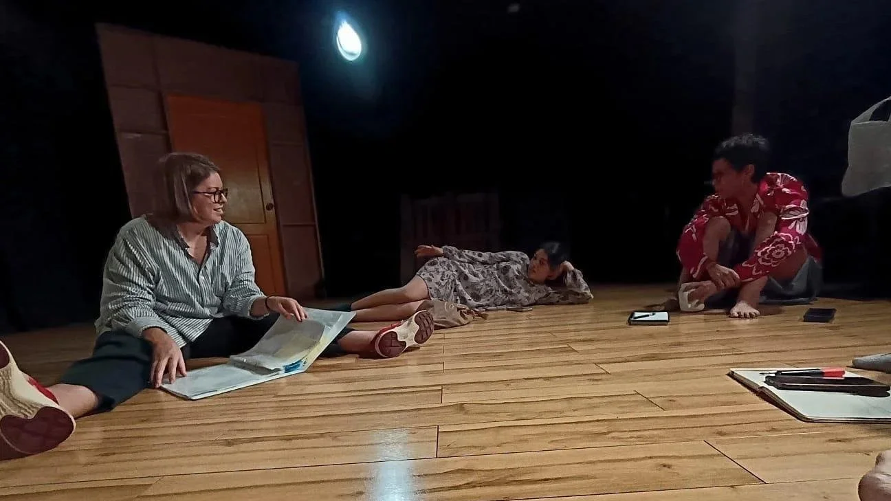 Three people sitting and lying on a wooden floor in a dimly lit room, engaging in conversation, with papers, a phone, and a notebook nearby.