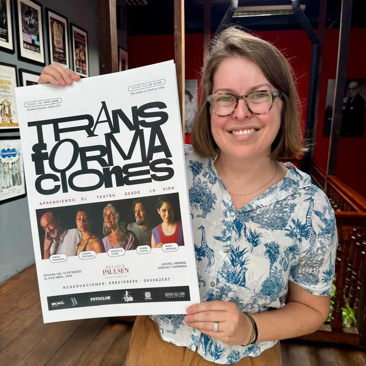 A woman wearing glasses and a blue and white patterned shirt holds a poster about a theatrical event titled "Transformaciones." She is smiling and standing indoors, with framed pictures on the wall behind her.