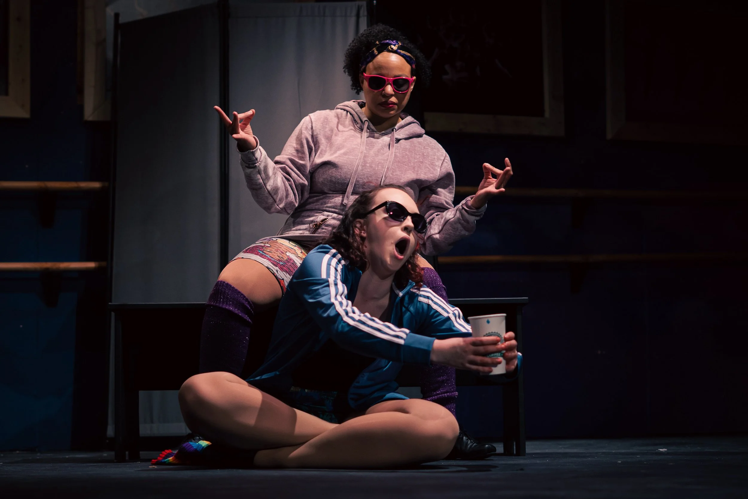 Two women in colorful outfits and sunglasses pose on a dark stage, one sitting on the floor holding a cup and the other standing behind her with arms raised, both with lively expressions.