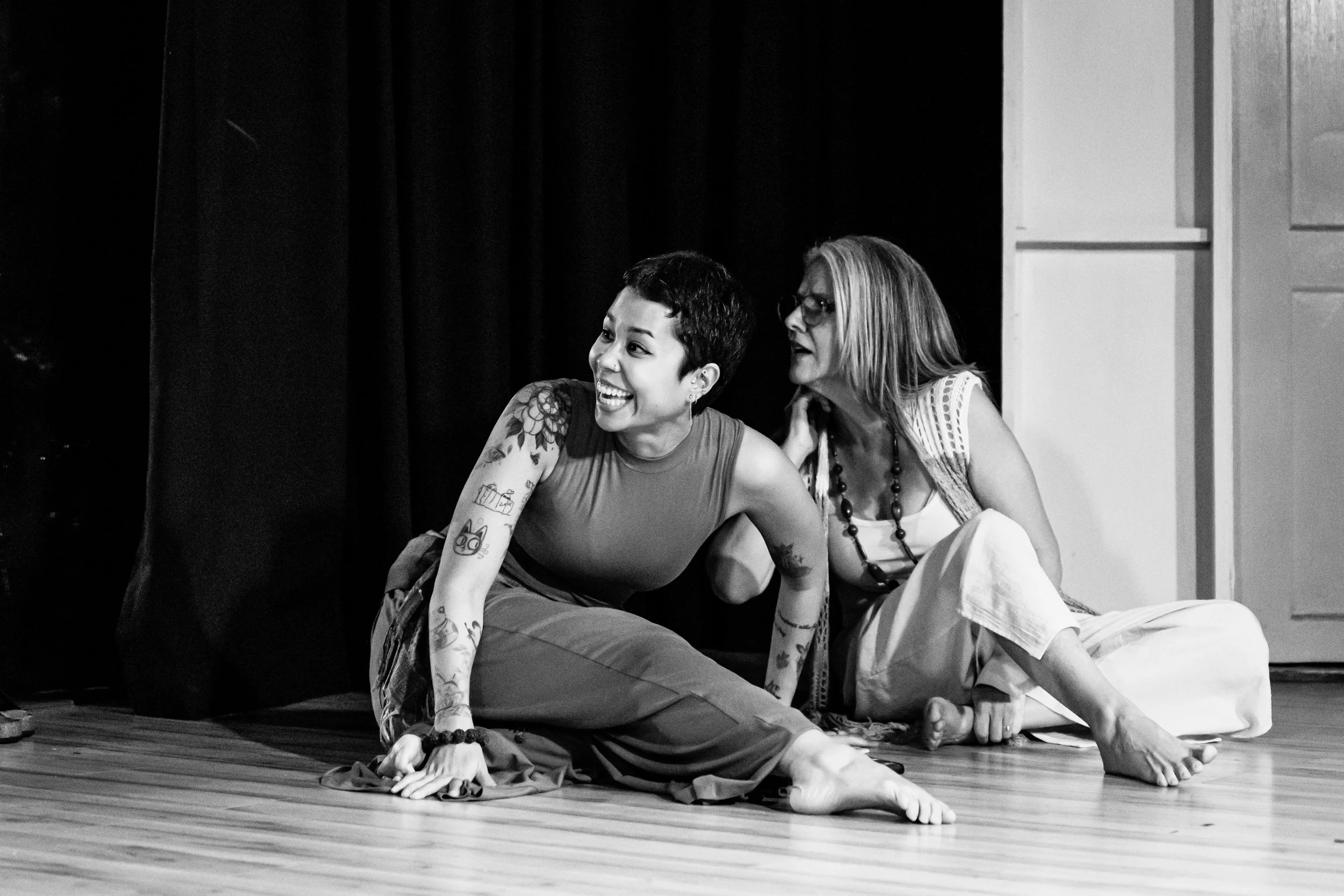 Two women sit on a wooden floor, one with tattoos and short hair smiling, the other with glasses and long hair looking at her, in front of a black curtain.