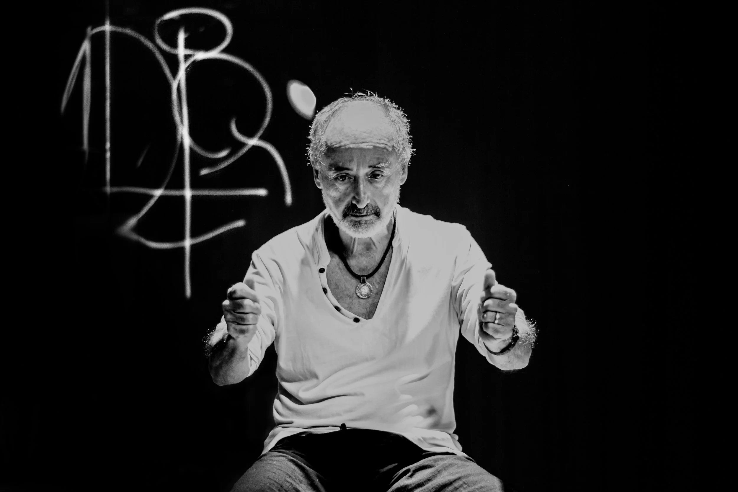 A black-and-white photo of an elderly man with gray hair and a beard, wearing a light-colored shirt with black buttons, clasping his fists and looking serious, with a dark background and white abstract lines on the left side.