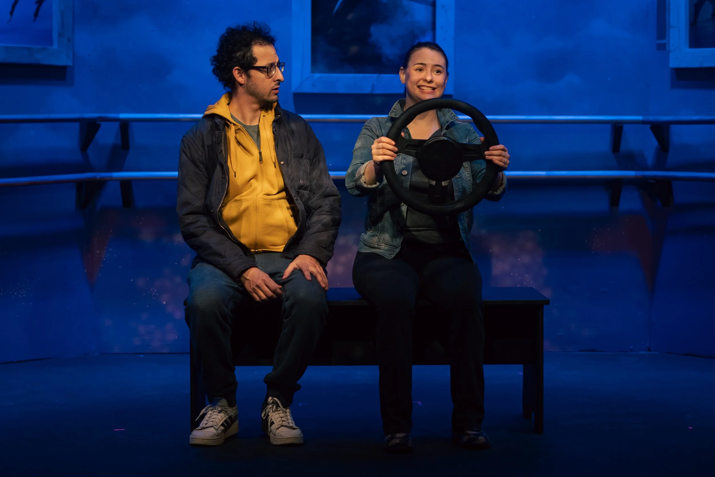 A woman holding a steering wheel and smiling, sitting next to a man with glasses, in front of a blue-lit stage or set, with empty benches behind them.