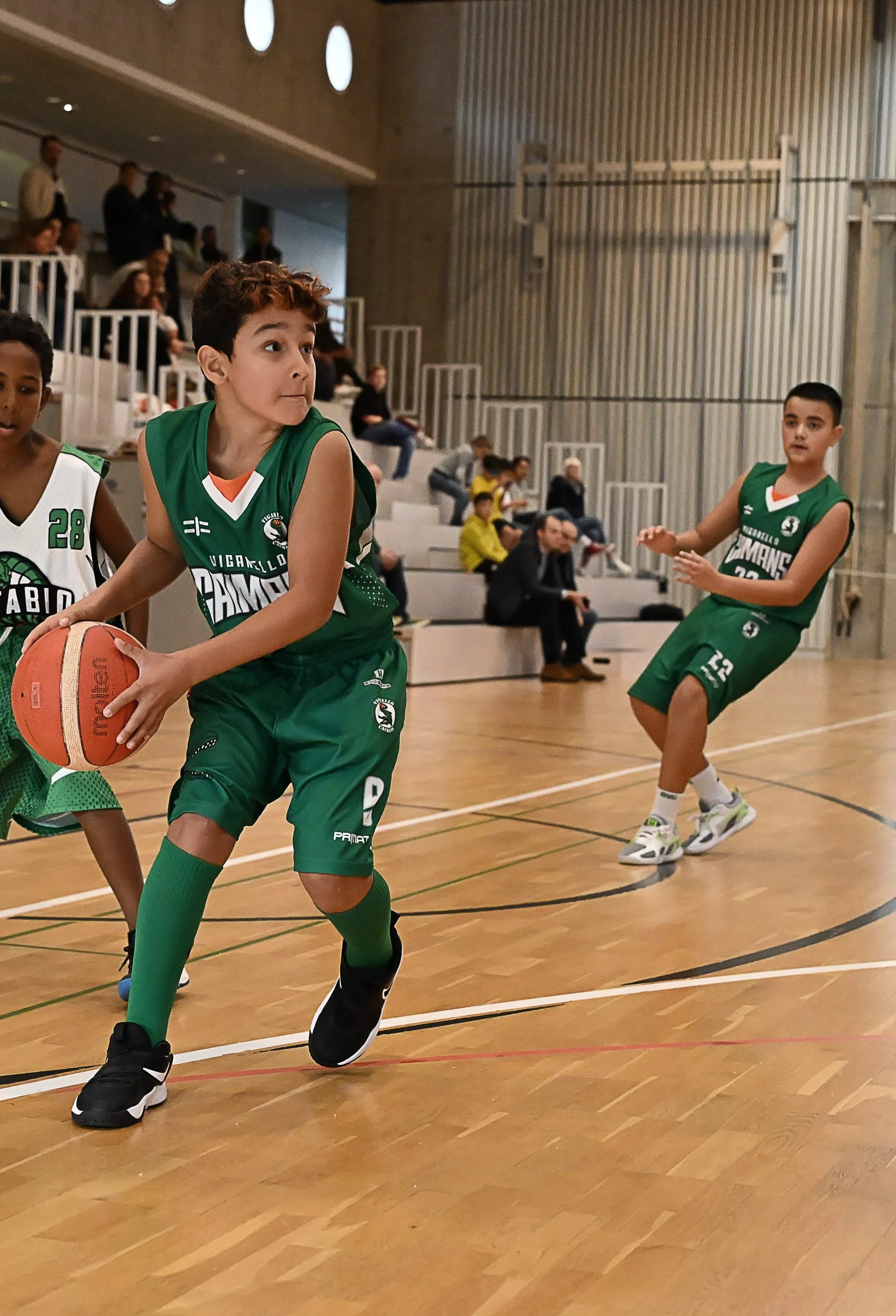 Giocatori di basket in azione durante una partita in un'arena indoor, con pubblico ad osservare sugli spalti.