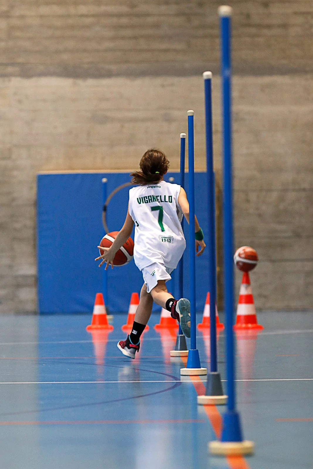 Bambina che corre e dribbla in un allenamento di basket indoor, con palloni e coni di traffico.