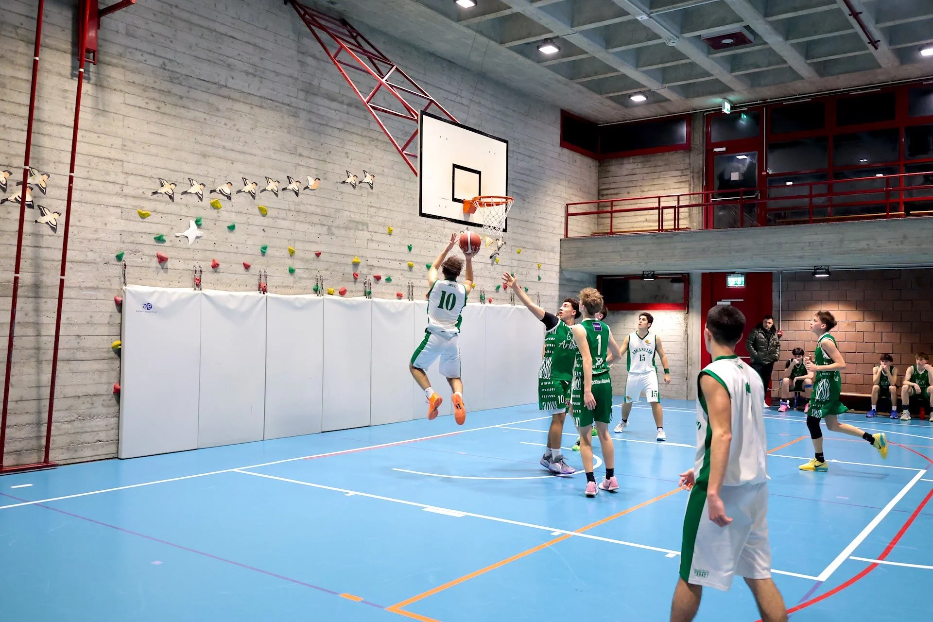 Giocatori di basket in azione in una palestra con pareti di cemento e parete da arrampicata e calcio a palla, alcuni seduti e alcuni in campo.