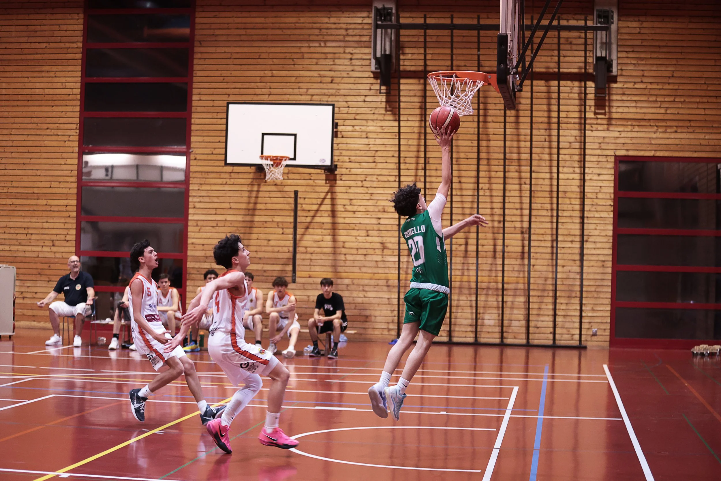 Giocatori di basket in azione durante una partita in palestra, con pubblico e arbitro sullo sfondo.