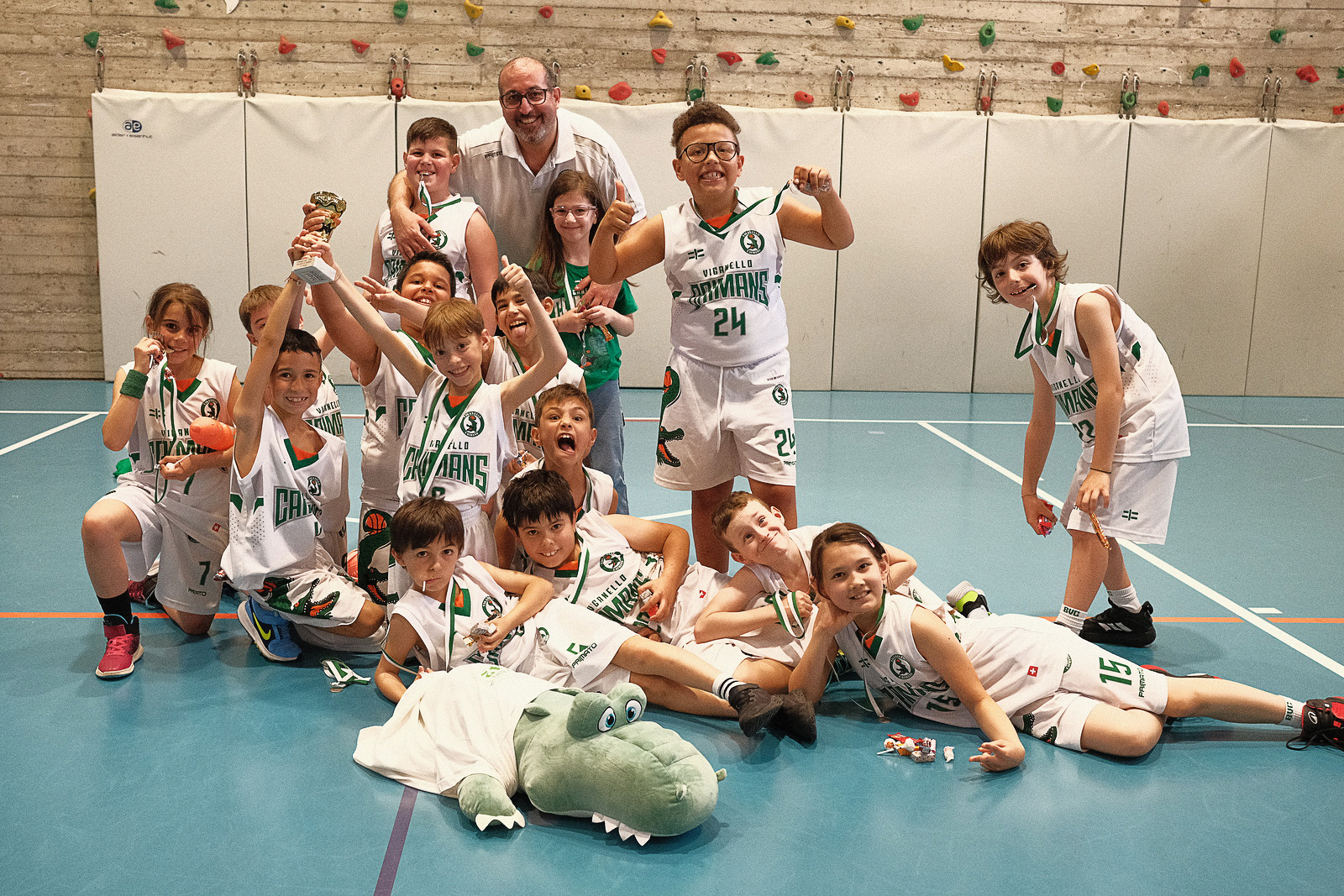 Gruppo di giovani giocatori di pallacanestro in uniforme bianca con medaglie, festeggiano una vittoria in palestra con un coach adulto dietro, alcuni seduti sul pavimento e altri in piedi, con un dinosauro gonfiabile e gadget fitness sul pavimento.