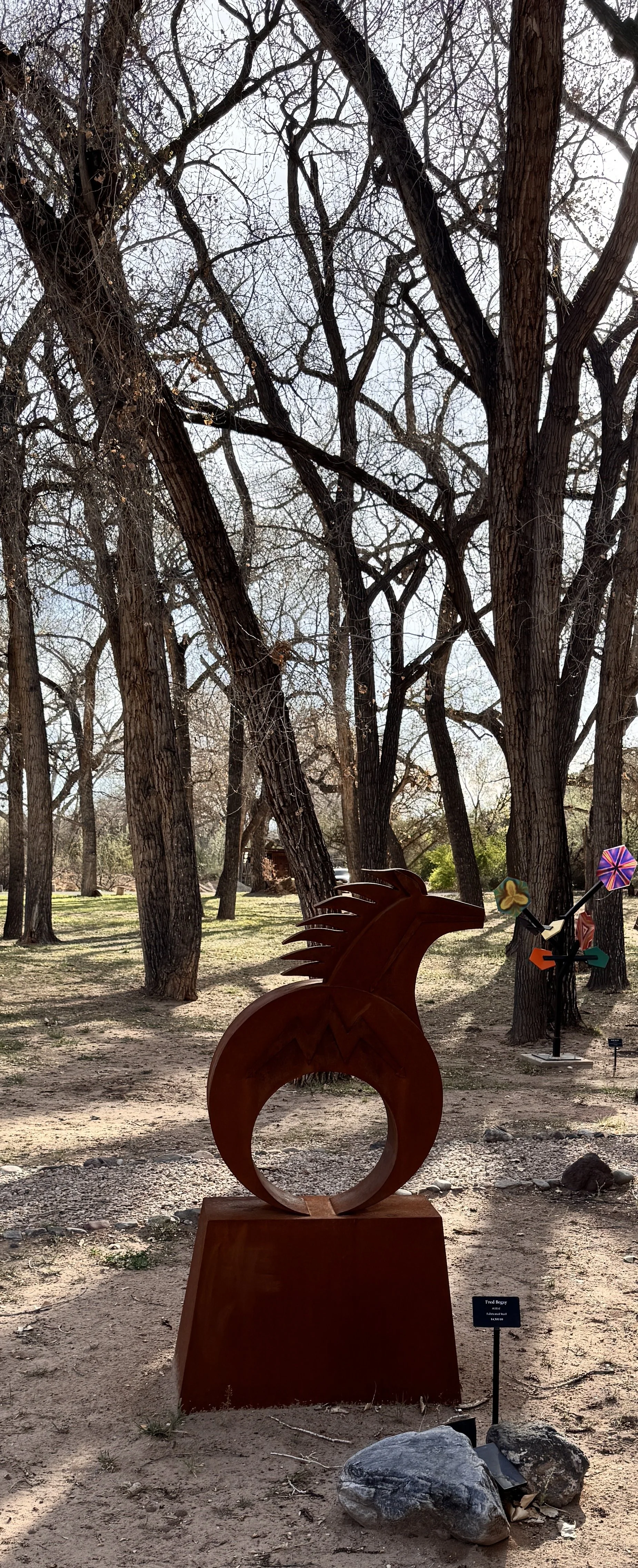 Outdoor scene with leafless trees and modern sculptures, including a large circular sculpture with a spiky design in the foreground and a colorful windmill sculpture in the background.