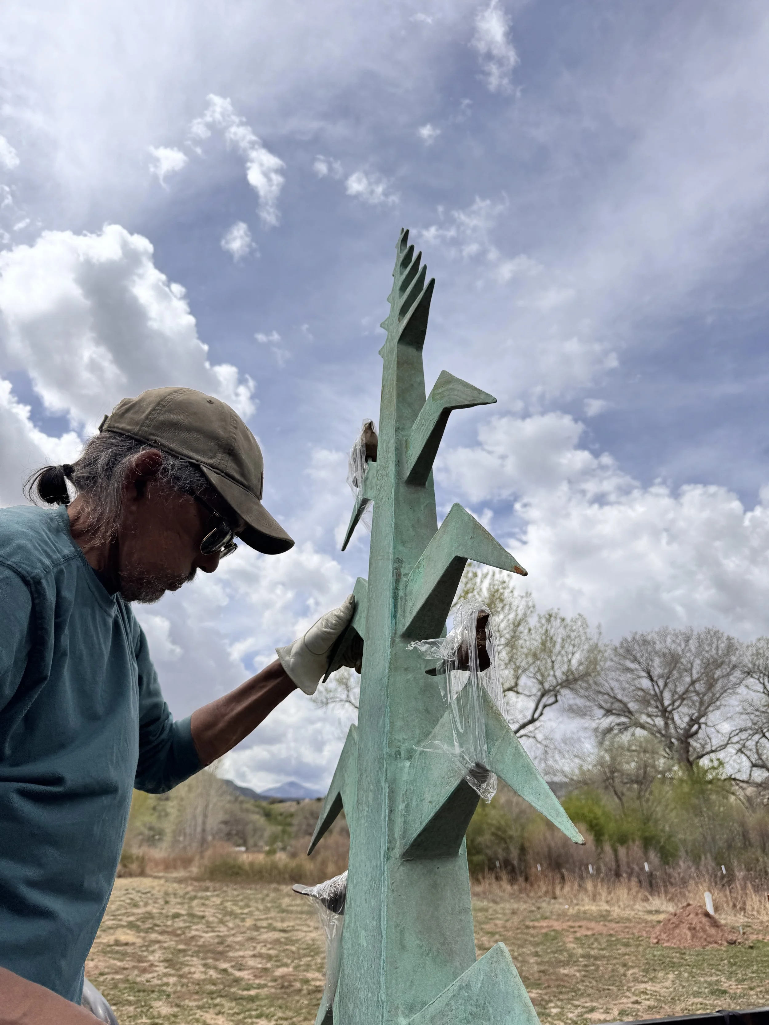 A person with gray hair, wearing a beige baseball cap, sunglasses, a blue long-sleeve shirt, and white gloves, working on a tall outdoor metal sculpture shaped like a stylized tree or plant, with multiple pointed leaves or petals, under a partly cloudy sky with trees and mountains in the background.